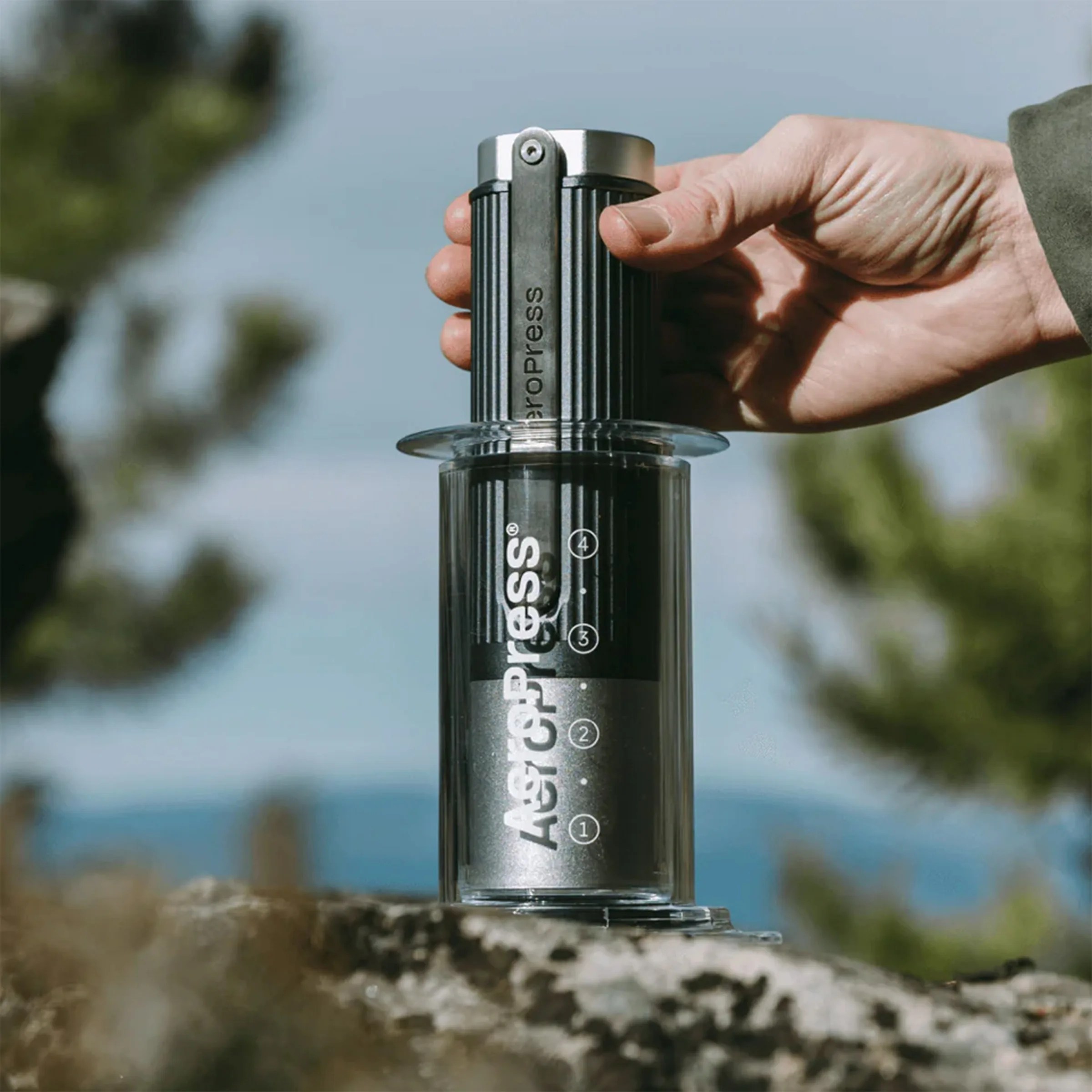 Hand holding a black and clear AeroPress coffee maker on a rock outdoors.