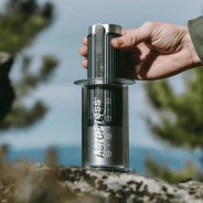 Hand holding a black and clear AeroPress coffee maker on a rock outdoors.