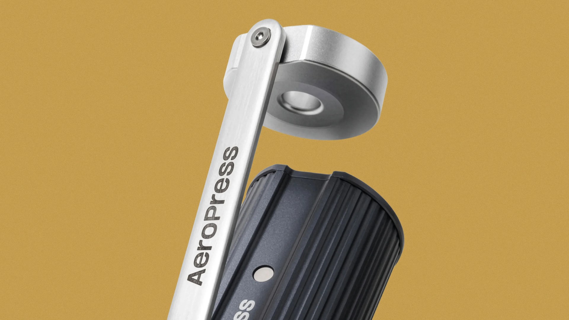 Close-up of a black AeroPress coffee maker with a silver metal press arm against a mustard yellow background.