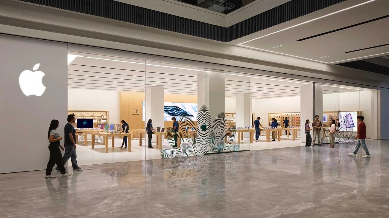 Apple store with glass walls, wooden tables displaying products, and people walking and shopping inside.