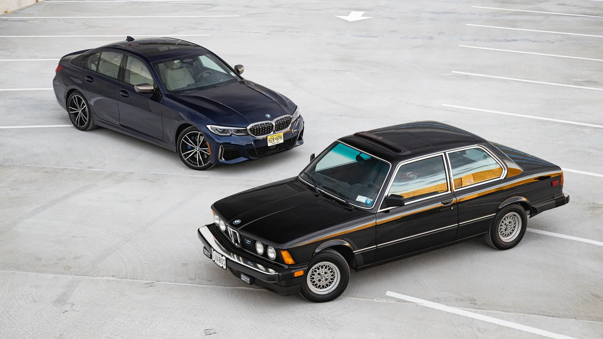 Two BMW sedans, one black vintage model with chrome trim and one dark blue modern model, parked on a concrete lot.