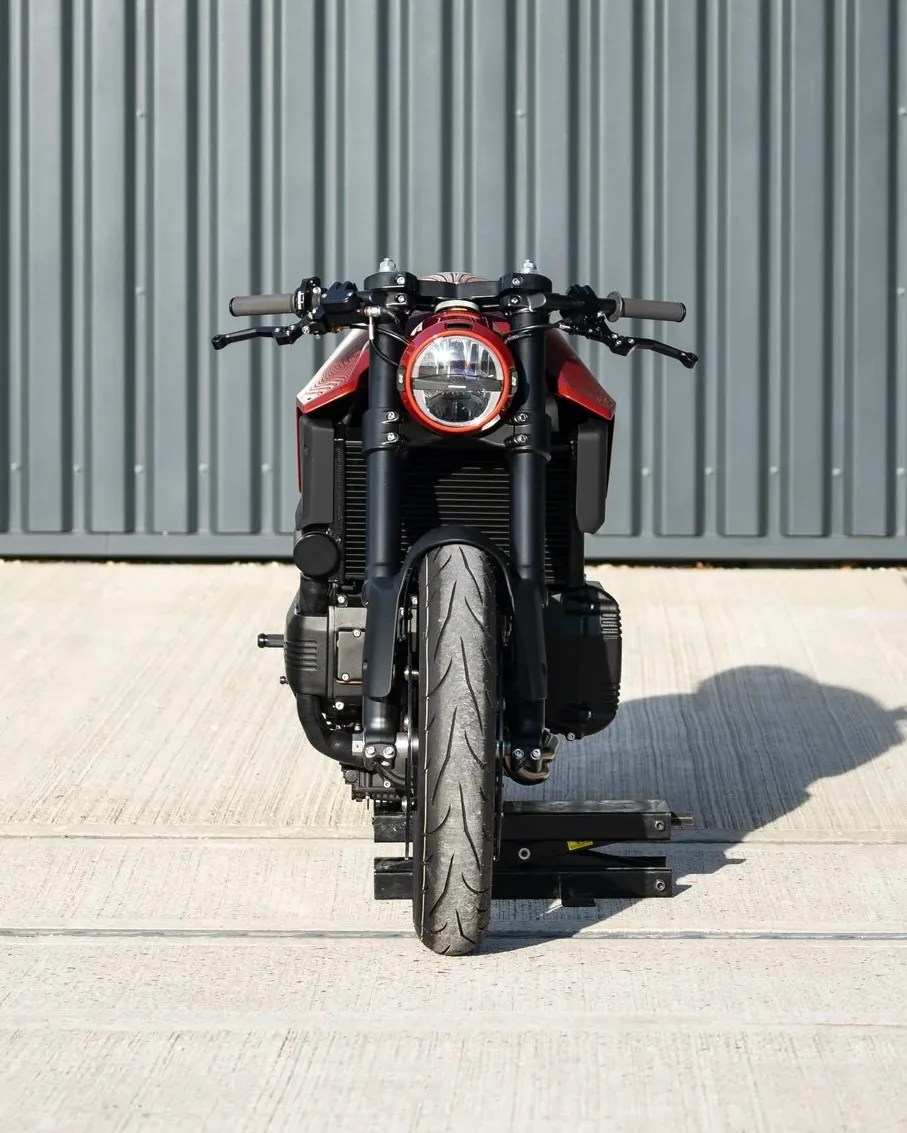 Front view of a black and red motorcycle with a round headlight, parked on a concrete surface in front of a corrugated metal wall.