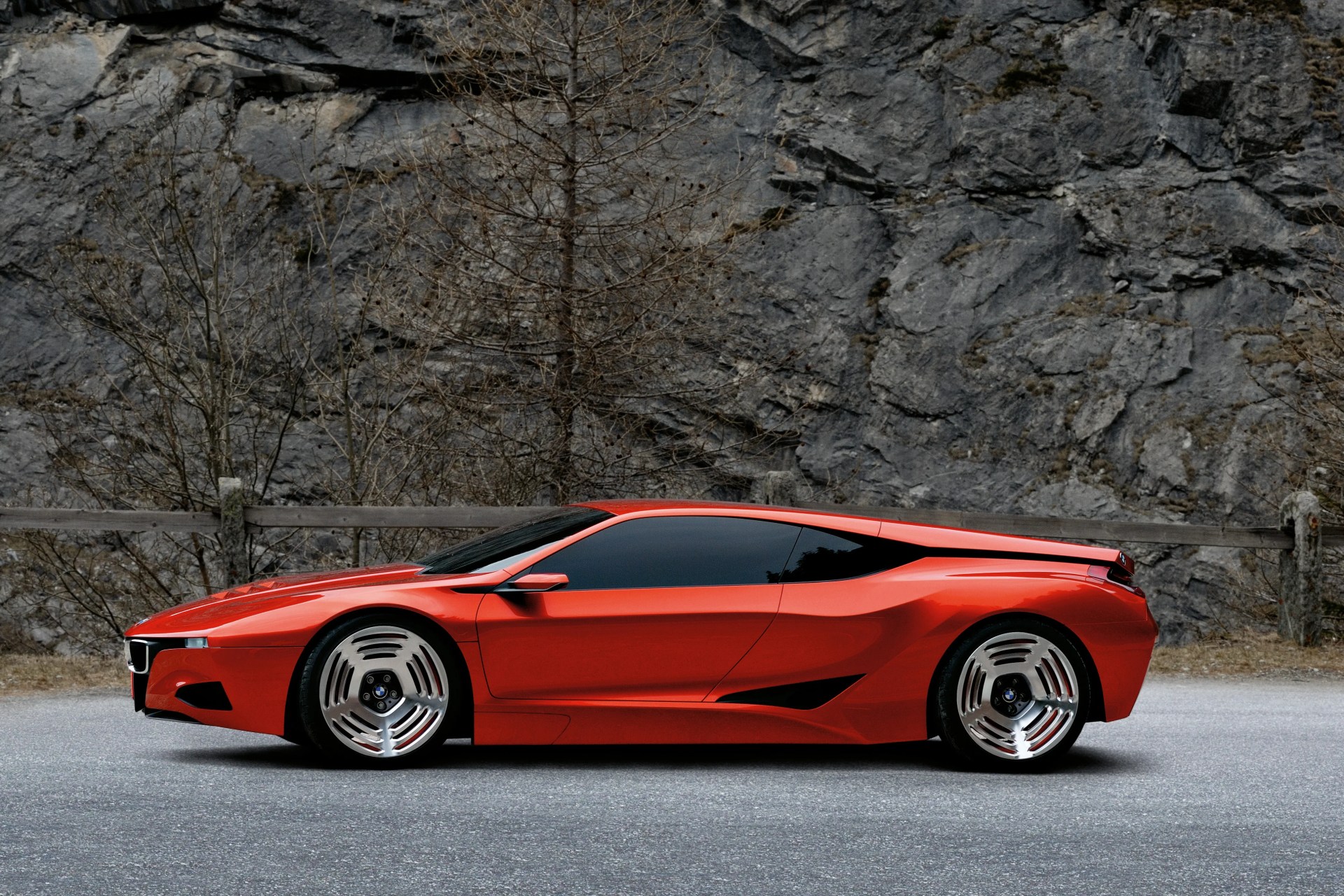 Sleek red sports car with tinted windows and unique silver alloy wheels parked on a road with rocky cliffs in the background.