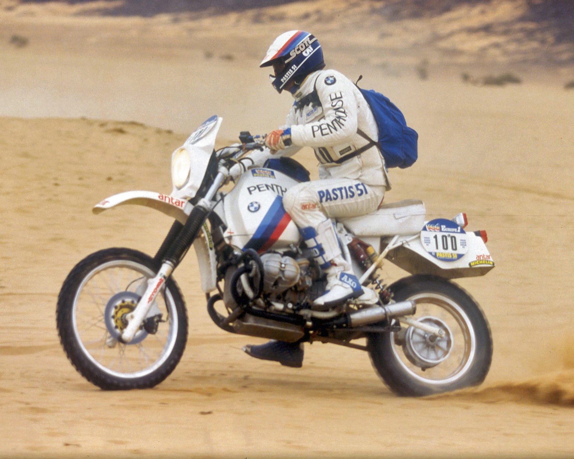 White BMW off-road motorcycle with racing decals ridden on sand by a person in white racing gear and blue backpack.