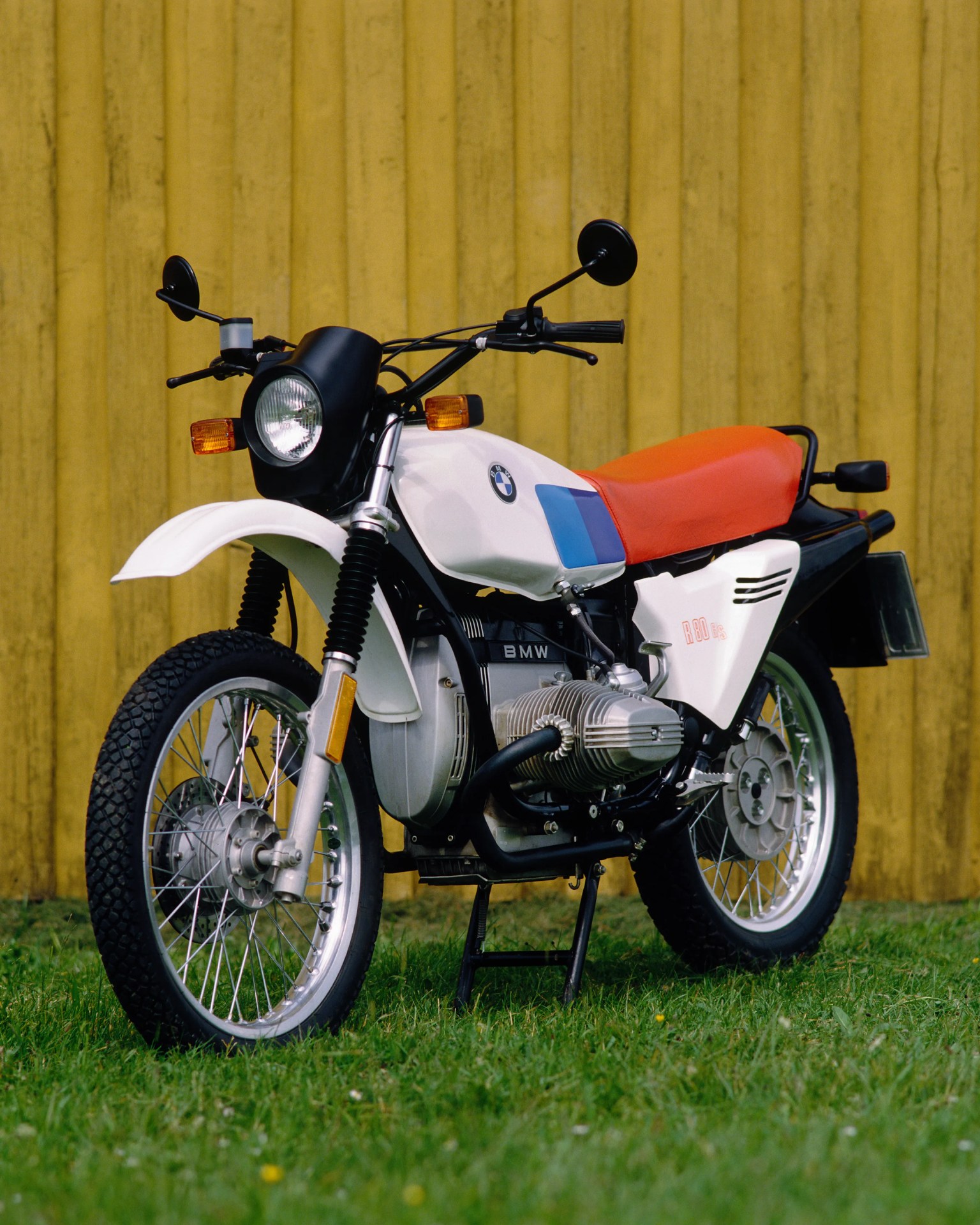 White BMW R80 GS motorcycle with orange seat parked on grass in front of a yellow wooden wall.