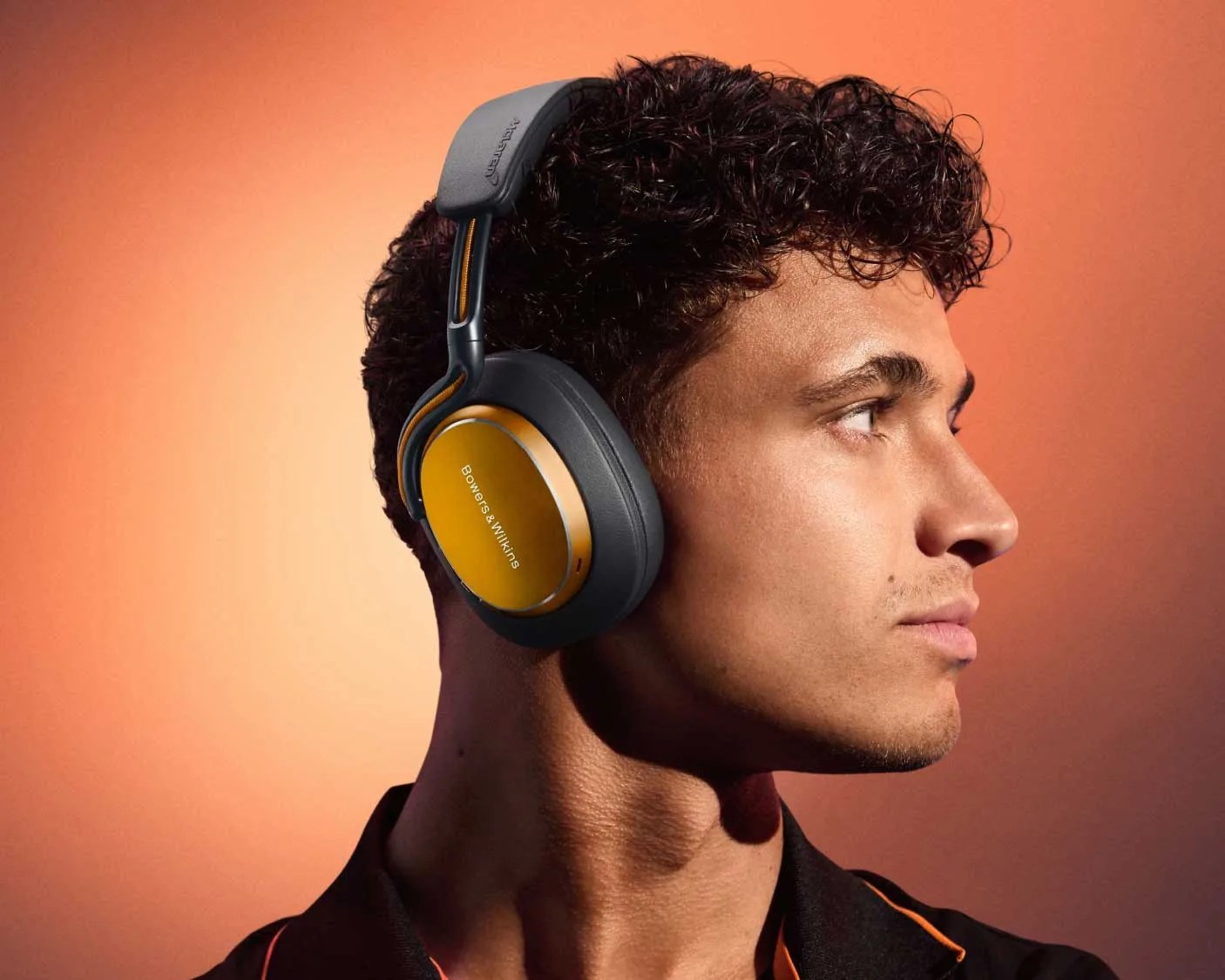 Person wearing black and orange over-ear Bowers & Wilkins headphones with gold ear cups against an orange background.