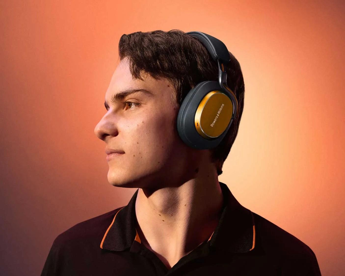Person wearing black and gold Bowers & Wilkins over-ear headphones against an orange background.