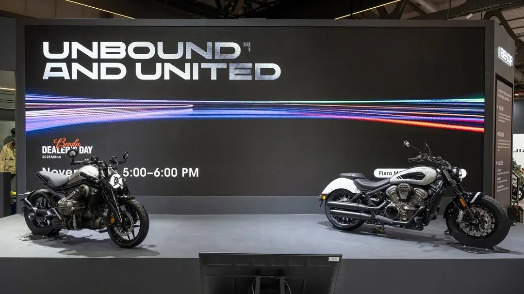 Two black and white motorcycles displayed on a stage with a large screen behind showing "UNBOUND AND UNITED" text.