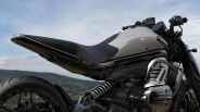 Close-up of a black and silver motorcycle seat, rear suspension, and engine against a blurred outdoor background.