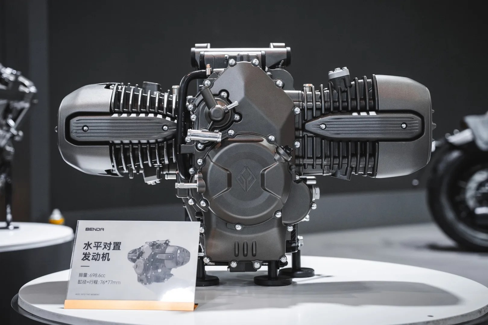 Horizontal twin-cylinder motorcycle engine with black casing displayed on a white round table.