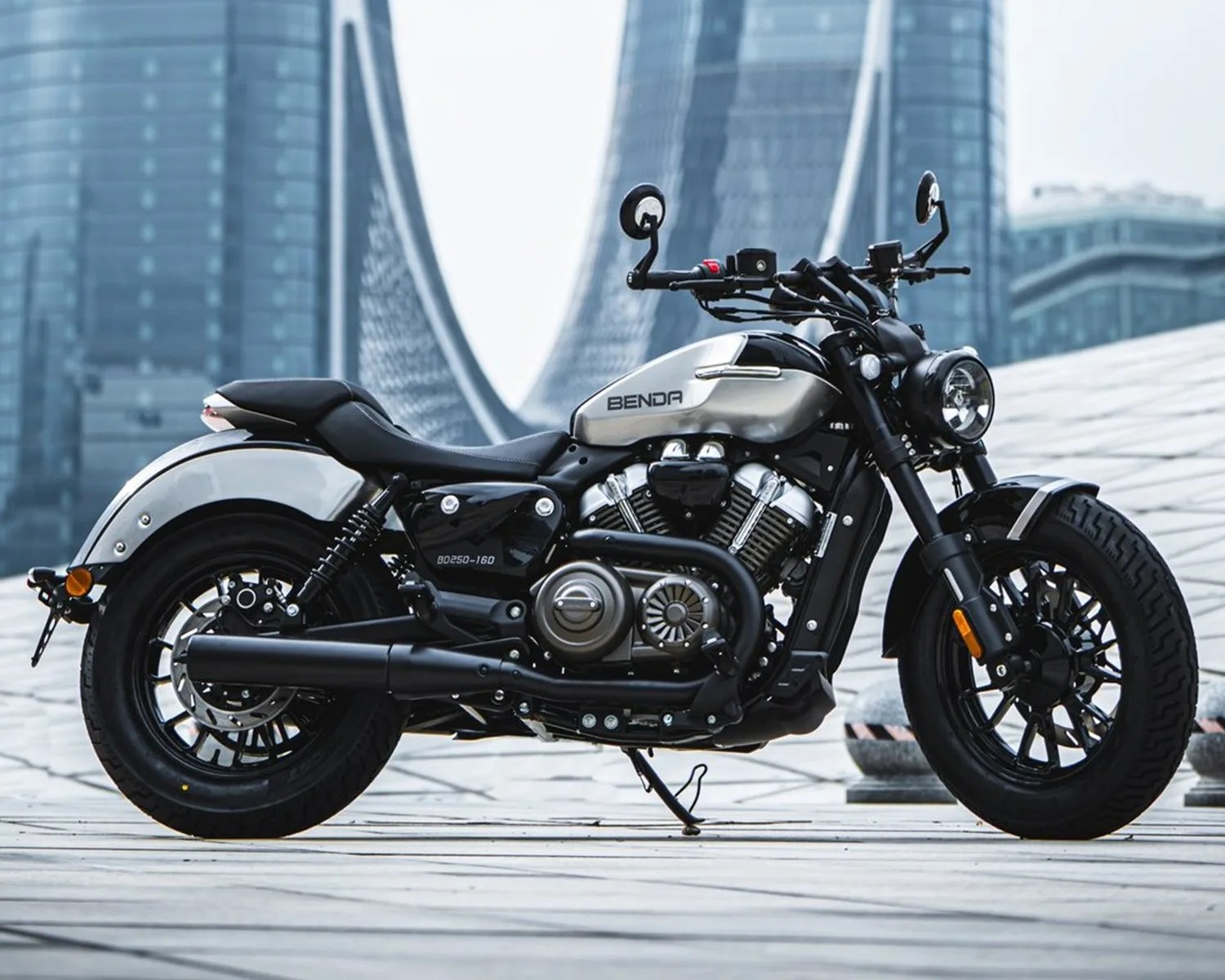 Silver and black Benda cruiser motorcycle parked on a tiled urban plaza with modern buildings in the background.