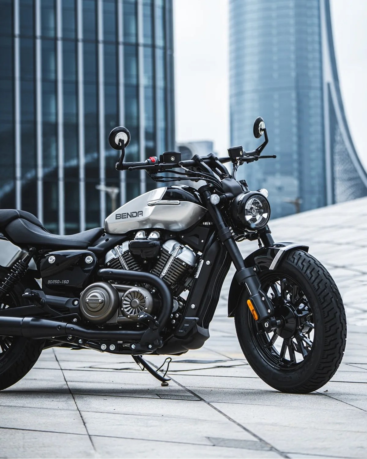 Silver and black Benda BD250-160 motorcycle parked on a tiled urban plaza with modern glass buildings in the background.