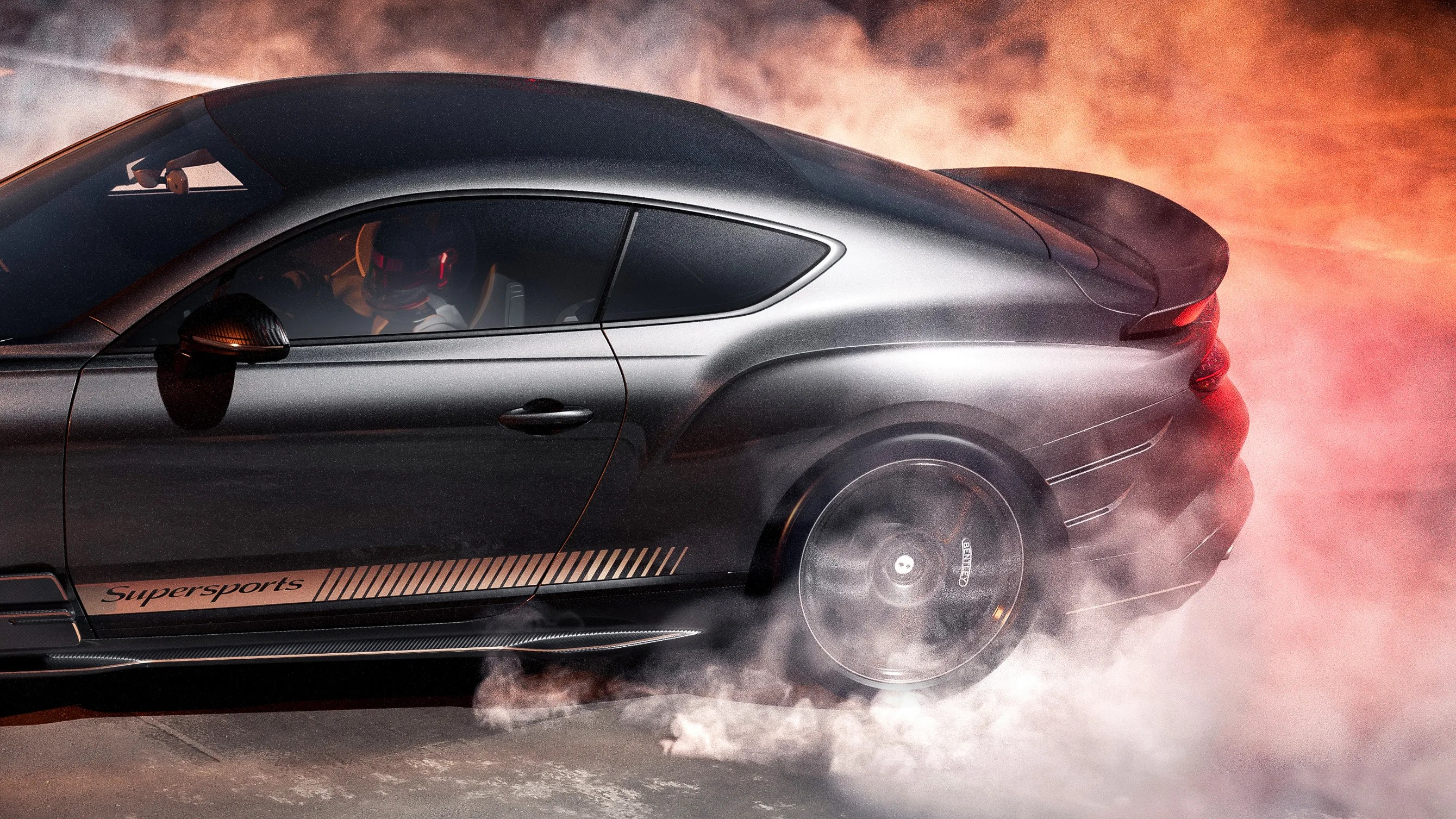 Side view of a dark gray Bentley Supersports coupe with rear tire spinning and smoke around the wheel.