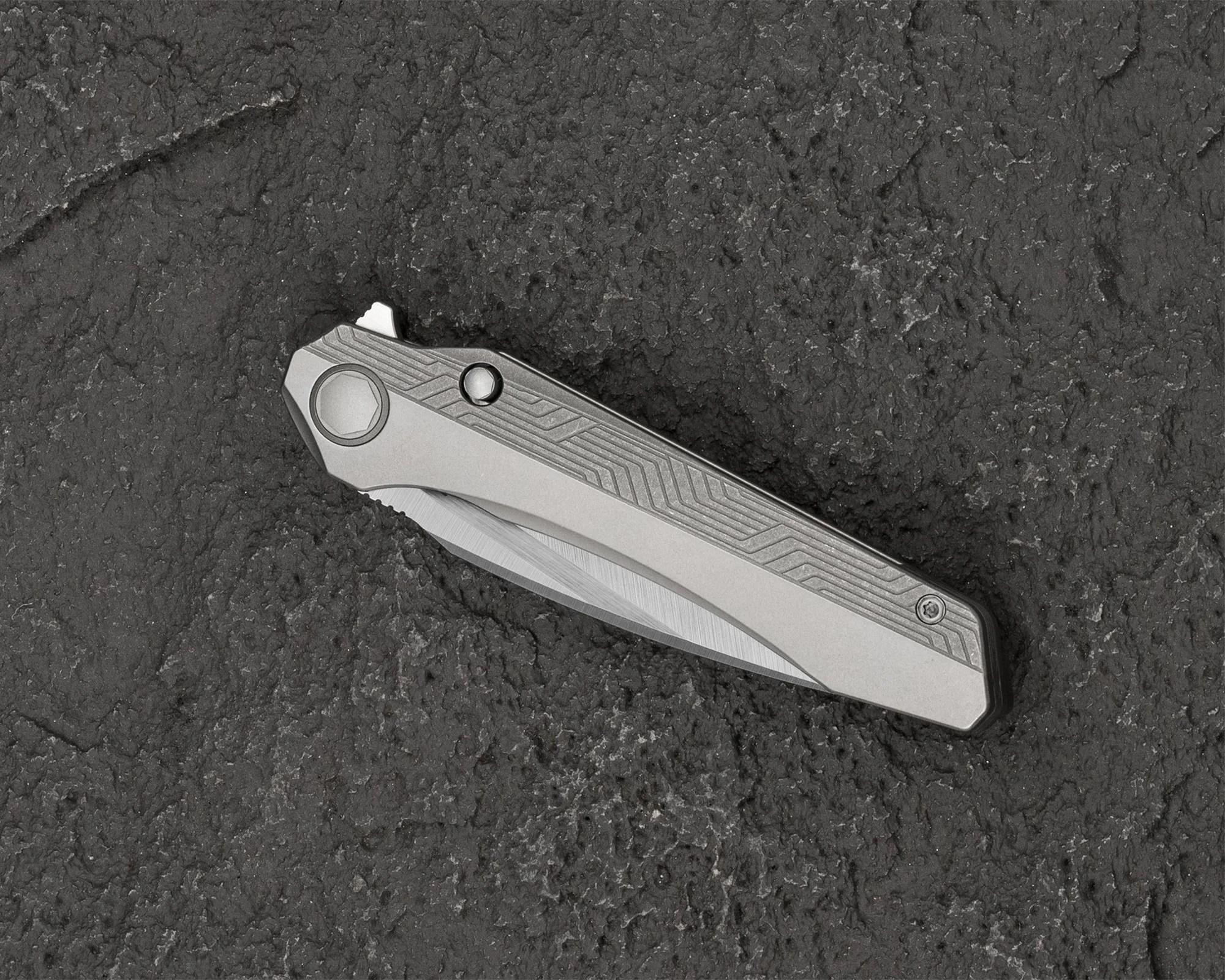Folded silver metal pocket knife with geometric patterns on the handle on a textured dark surface.