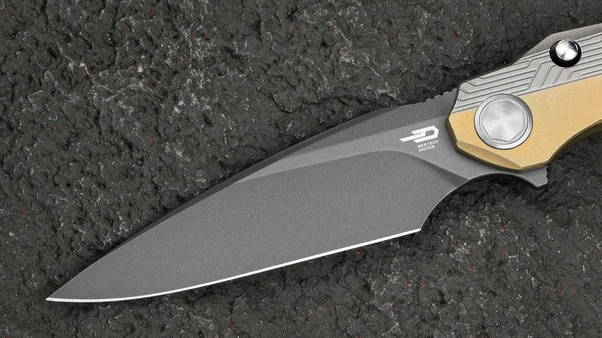 Gray blade with angular design and Bestech Knives logo, attached to a gold and silver handle on a rough black surface.