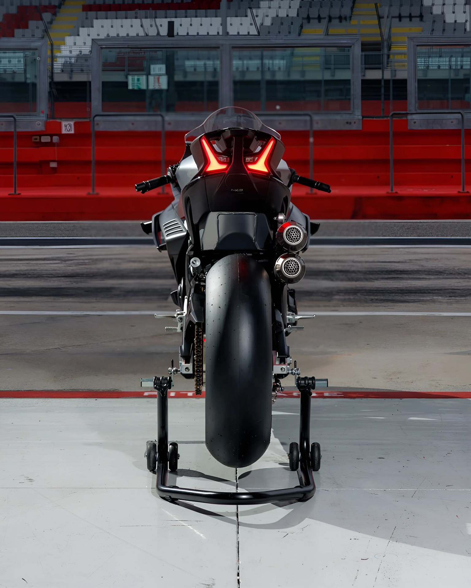 Rear view of a black racing motorcycle with dual exhausts on a stand in a racetrack pit lane.