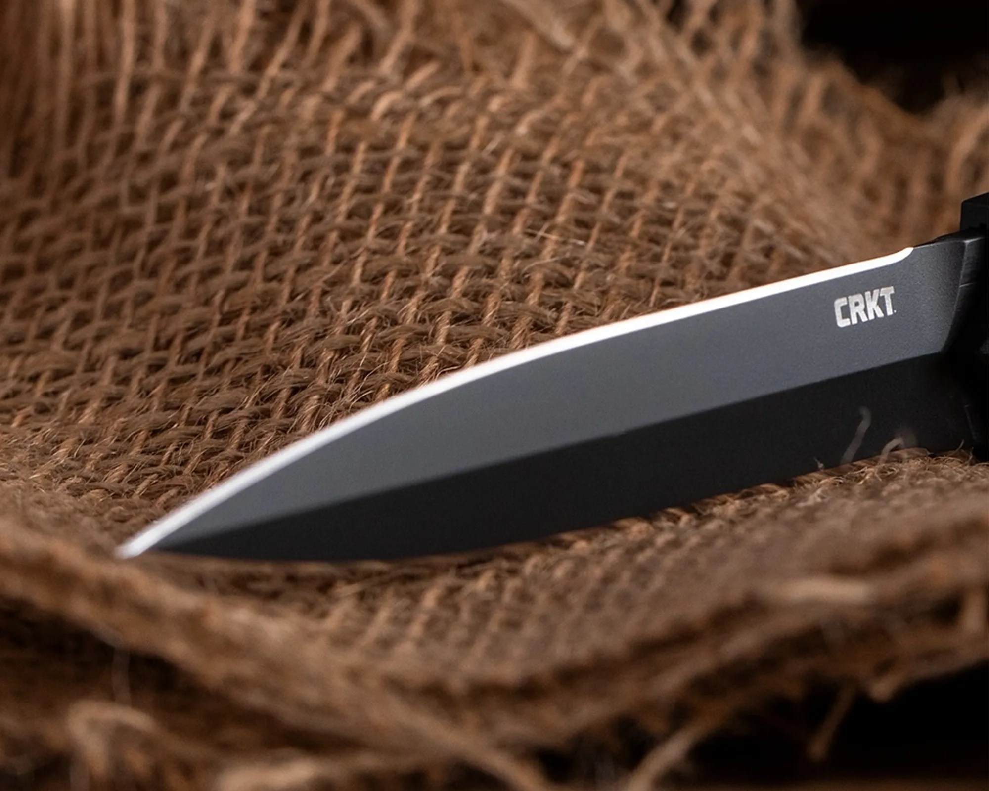 Close-up of a black CRKT knife blade resting on coarse brown burlap fabric.
