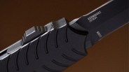 Close-up of a black folding knife handle with textured grip and partially visible blade marked "RODRIGUEZ DESIGN.