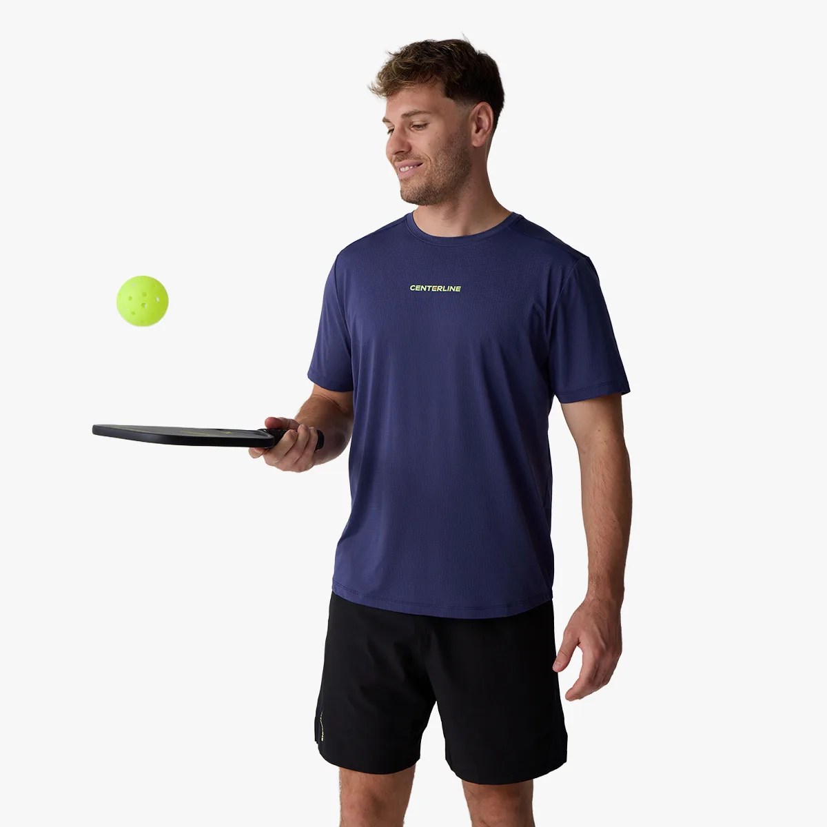 Man wearing a navy Centerline t-shirt and black shorts holding a black paddle with a yellow pickleball ball in the air.