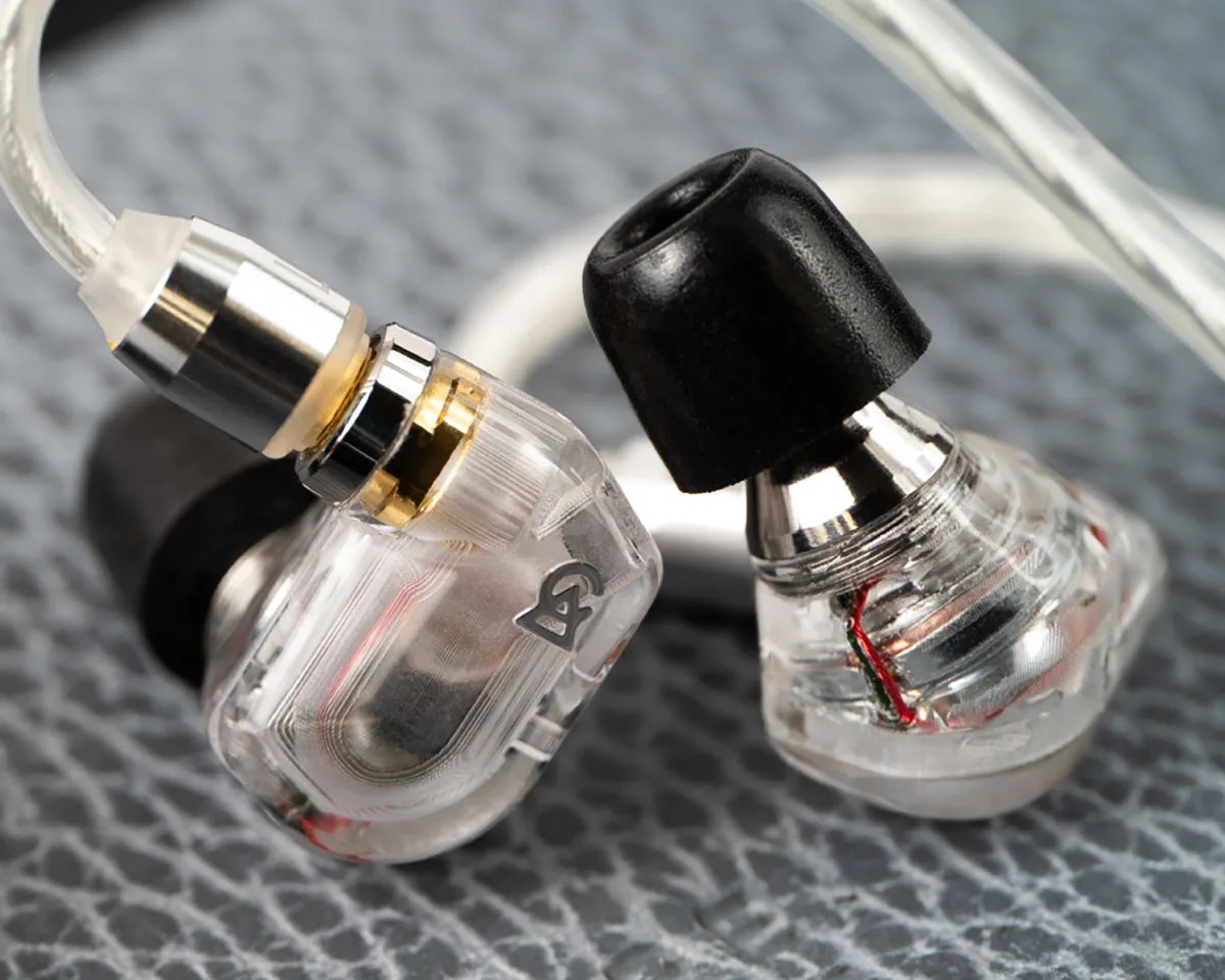 Pair of transparent in-ear monitors with black ear tips and visible internal wiring on a textured surface.