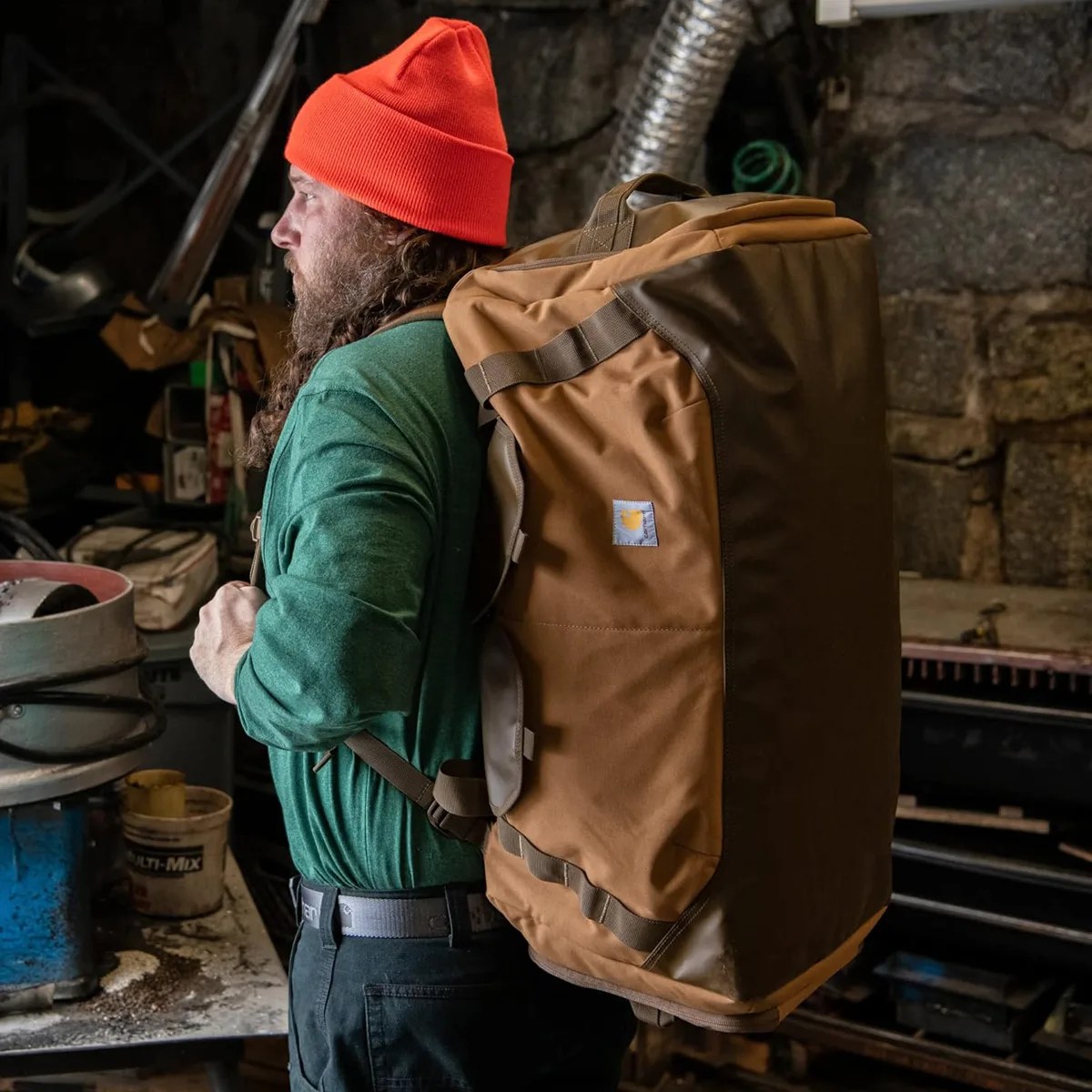Person wearing a large brown and dark brown backpack with a Carhartt logo, dressed in a green shirt and orange beanie.