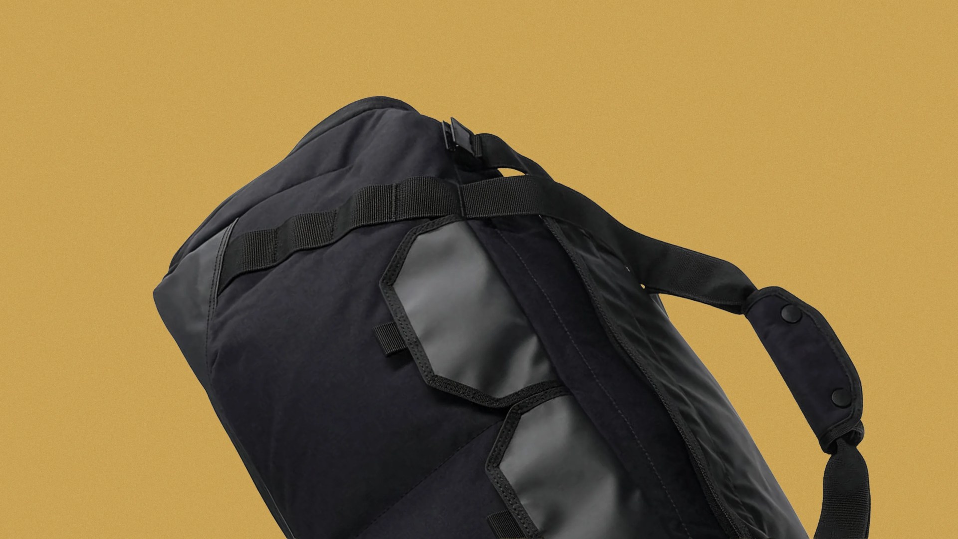 Black duffel bag with padded shoulder strap and webbing loops on a mustard background.