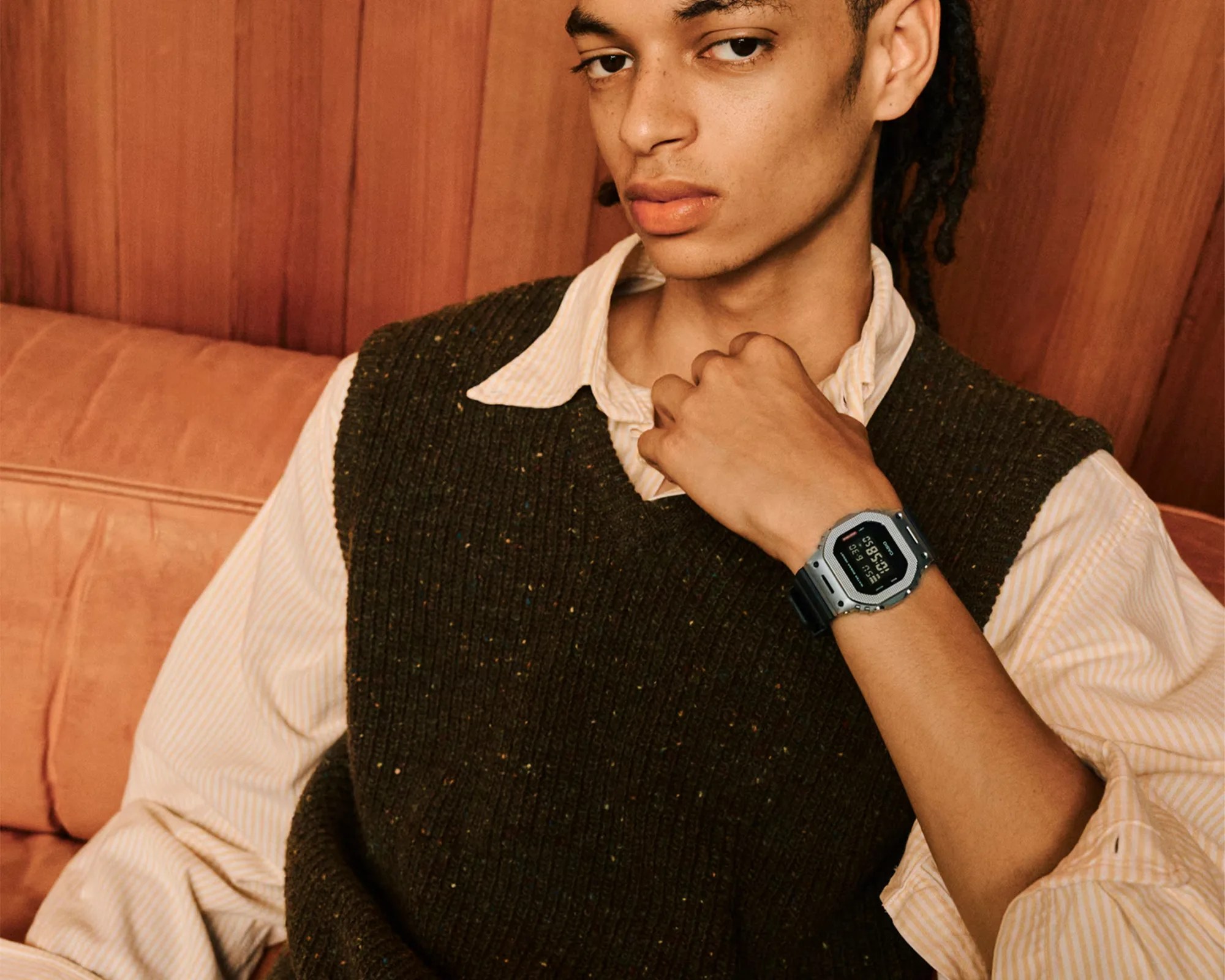 Person wearing a dark green knit vest over a light striped shirt, showing a silver digital wristwatch with a black strap.