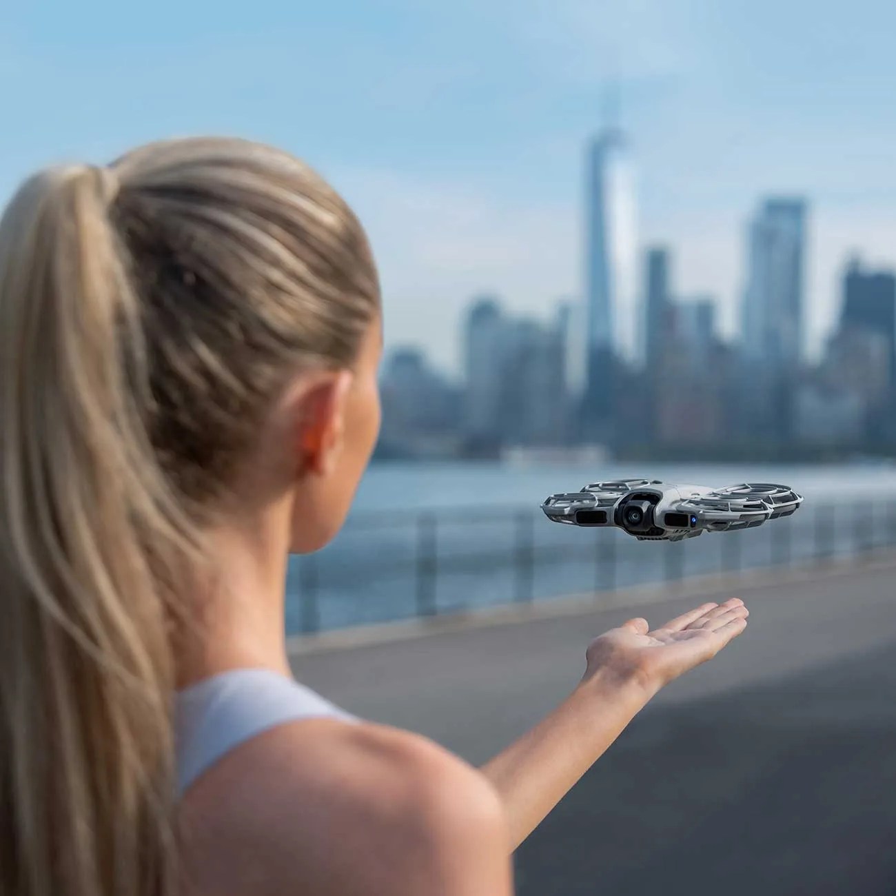 Woman with blonde ponytail holding a small gray drone hovering above her outstretched hand near a waterfront.