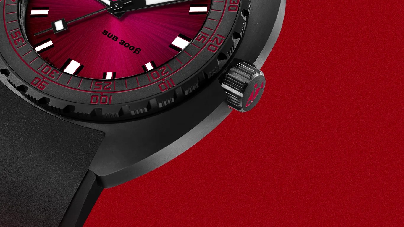 Close-up of a black dive watch with a red sunburst dial, black bezel with red markings, and black rubber strap on a red background.