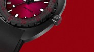 Close-up of a black dive watch with a red sunburst dial, black bezel with red markings, and black rubber strap on a red background.