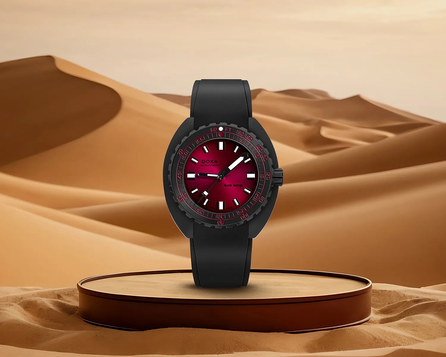 Black dive watch with a red gradient dial and black rubber strap displayed on a round platform in a desert setting.