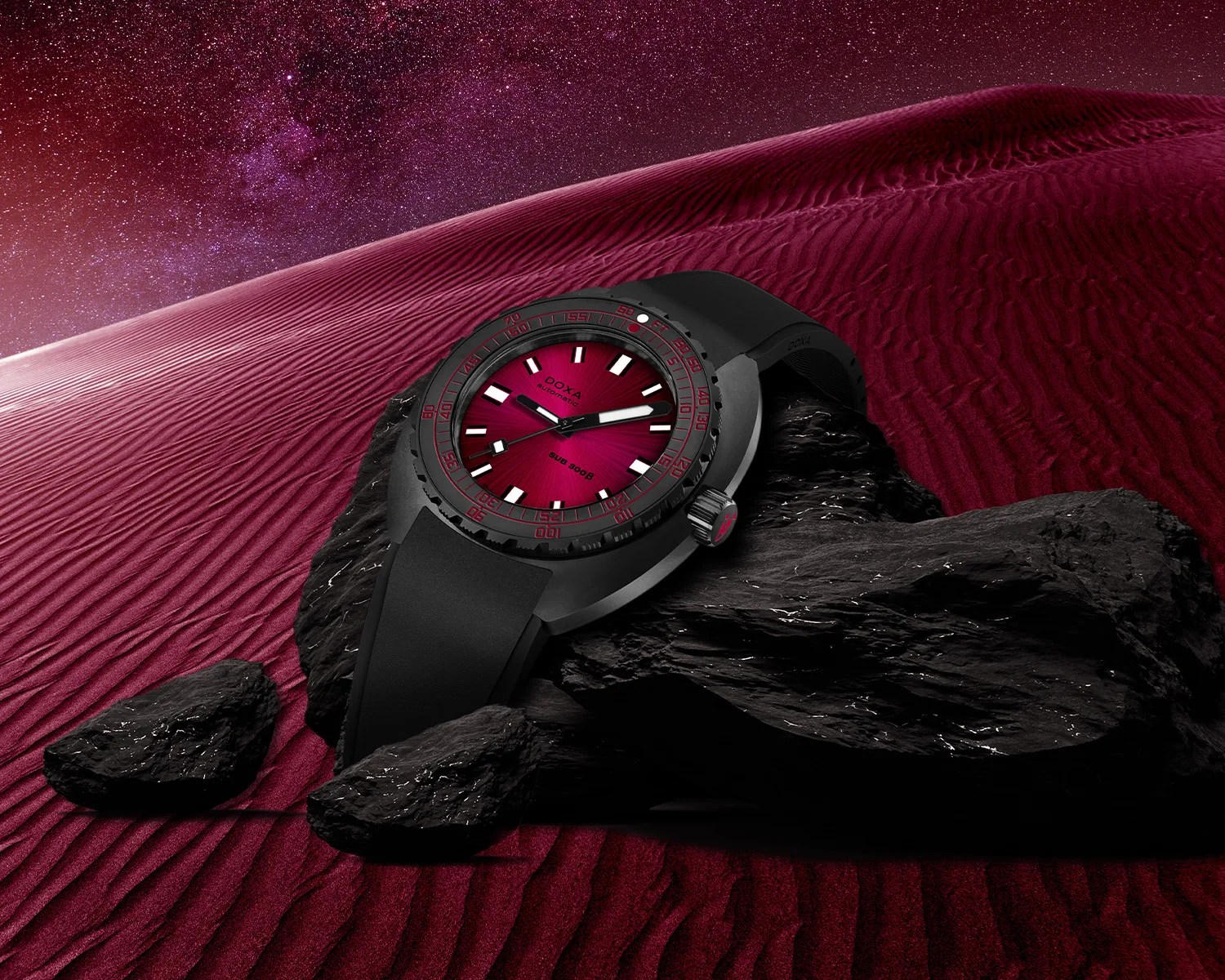 Black wristwatch with a red dial and black rubber strap resting on black rocks against a red sand dune and starry sky background.