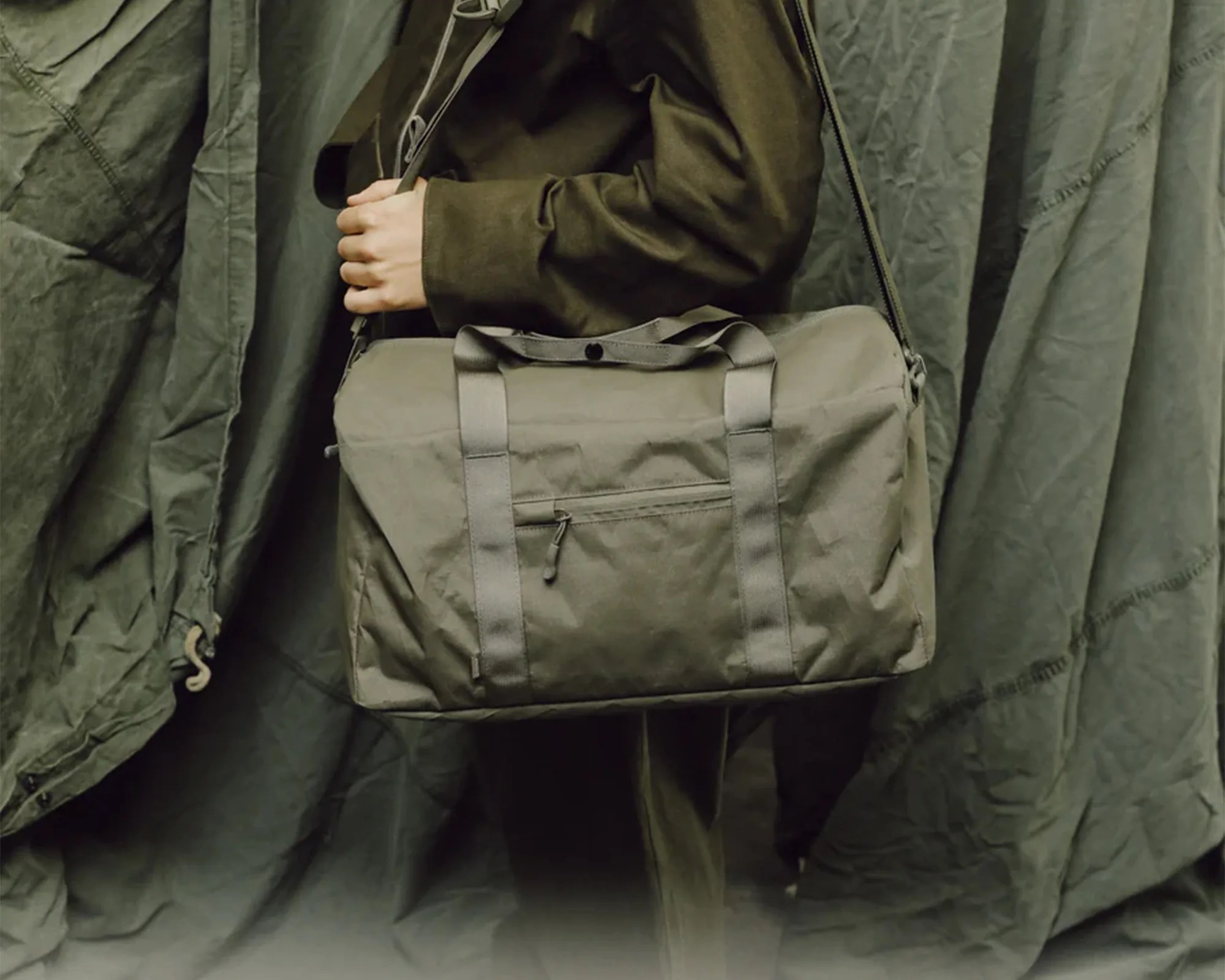 Olive-green canvas duffel bag with front zipper pocket worn crossbody by a person in matching green clothing.