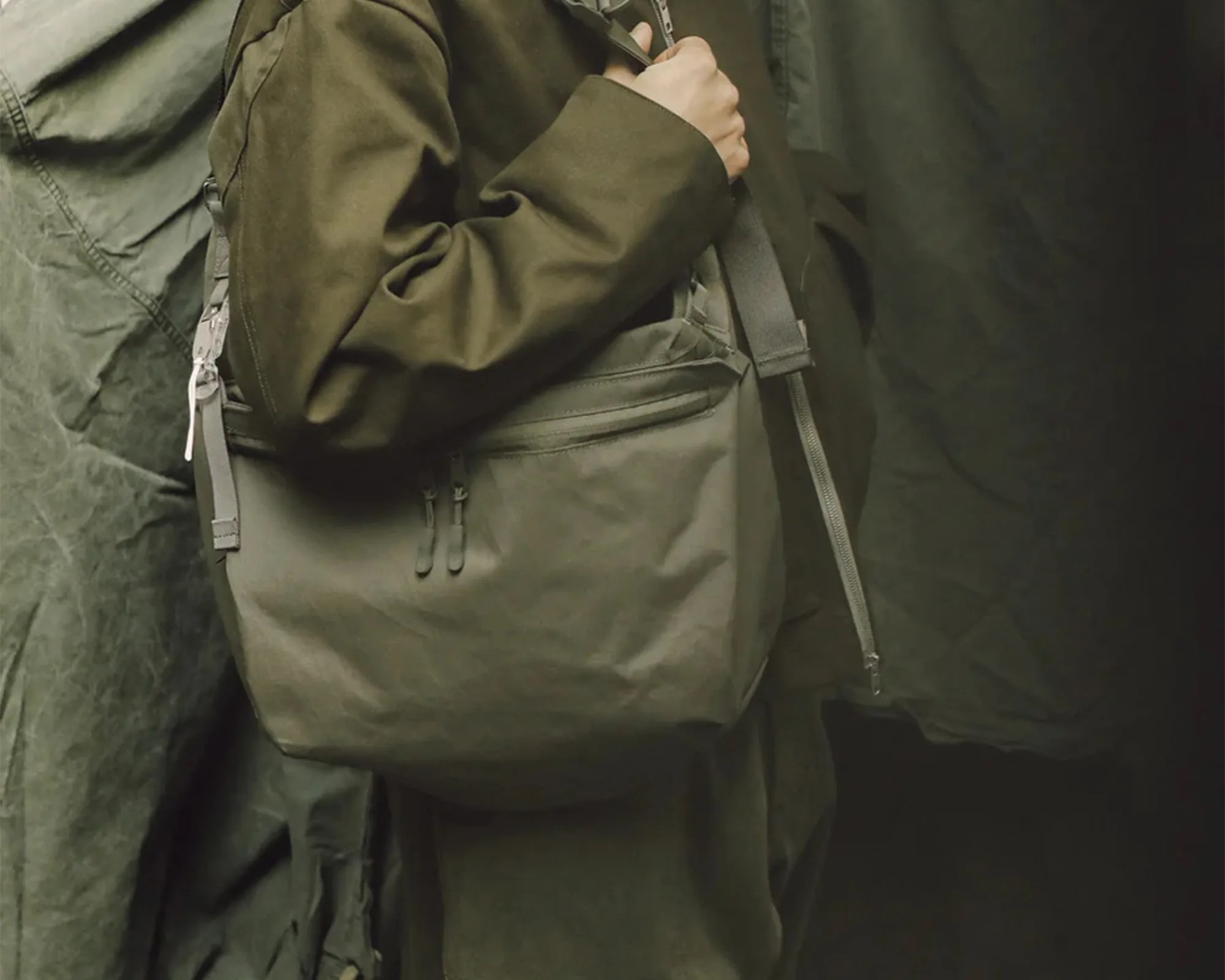Olive-green crossbody bag with dual zipper pockets worn over a matching olive-green jacket.