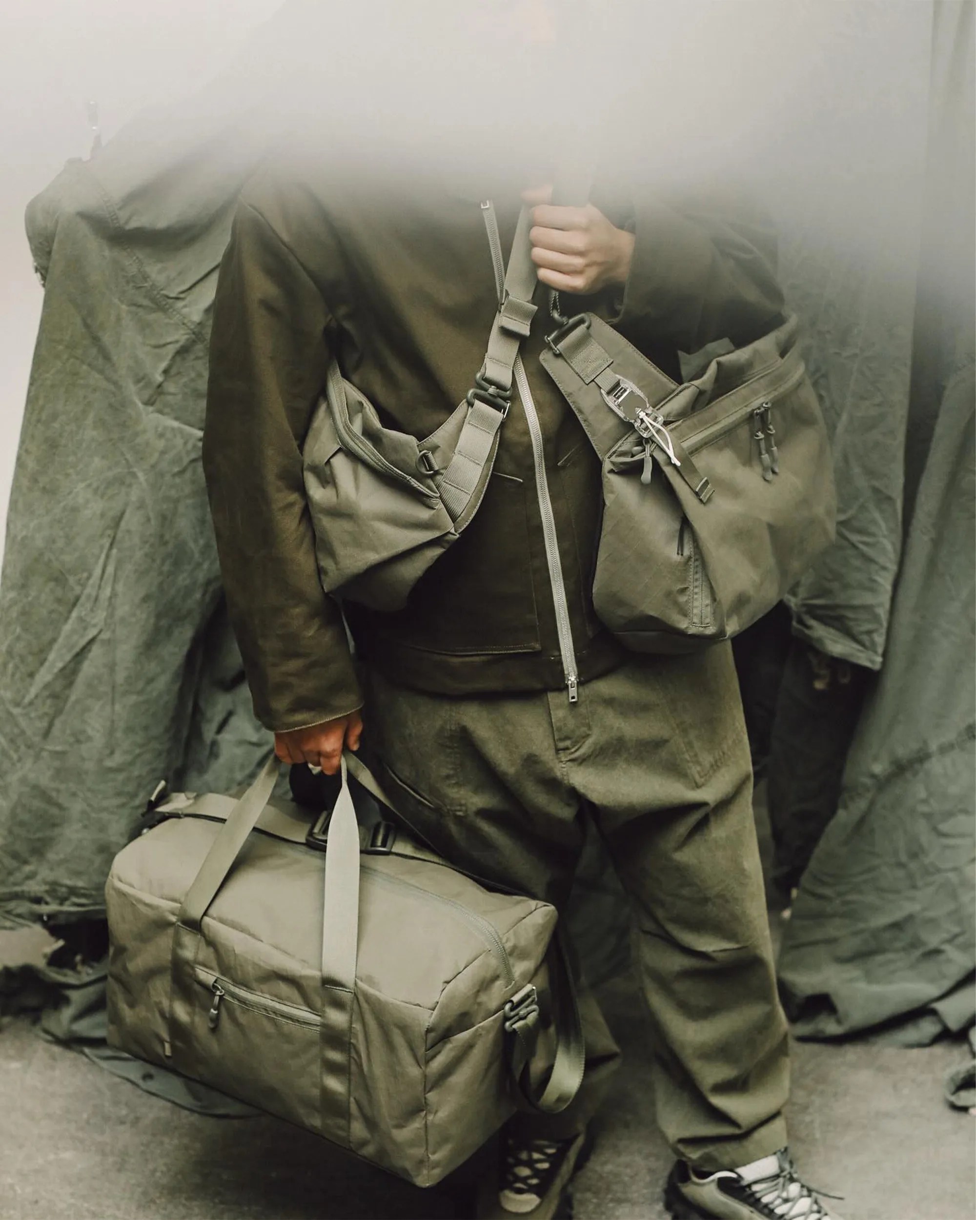 Person holding an olive-green duffel bag and wearing two olive-green crossbody bags with silver hardware.