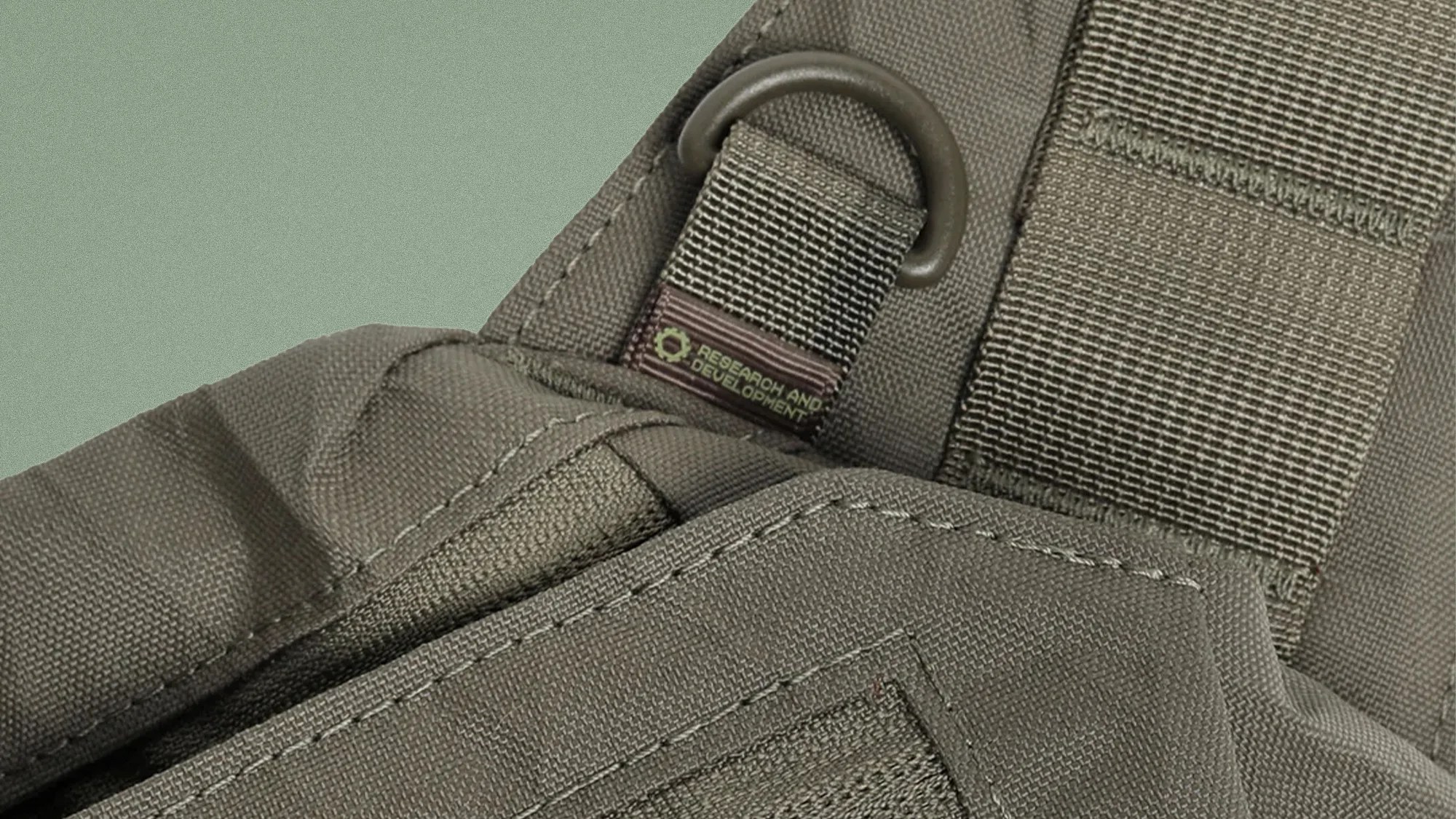 Close-up of olive green tactical fabric with a stitched strap and a metal D-ring labeled "Research and Development.