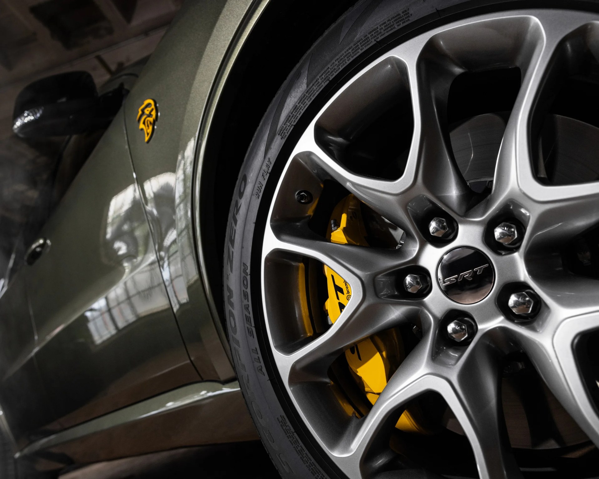 Close-up of a silver SRT alloy wheel with yellow Brembo brake caliper on an olive green car.