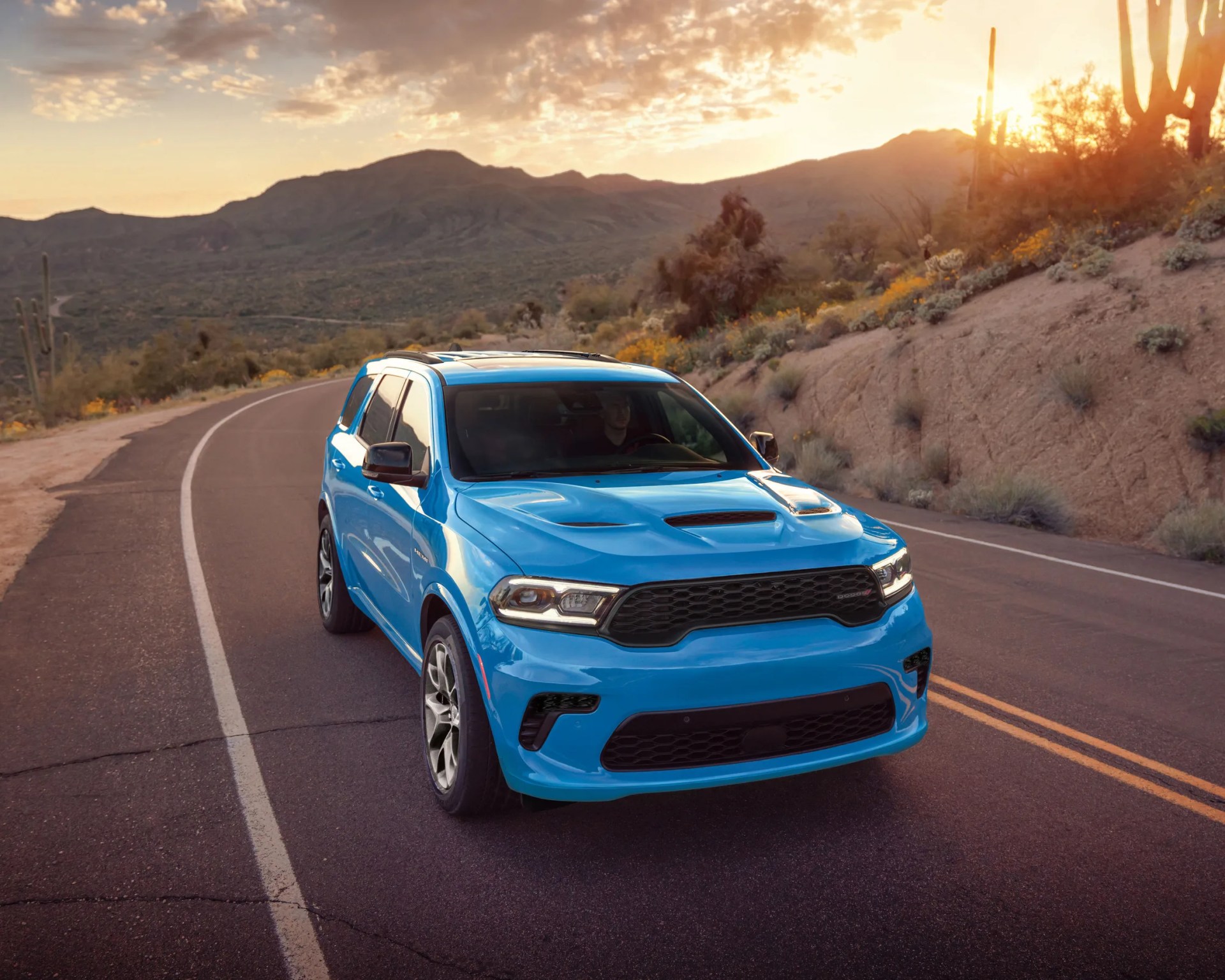 Bright blue Dodge Durango SUV on a winding desert road at sunset.