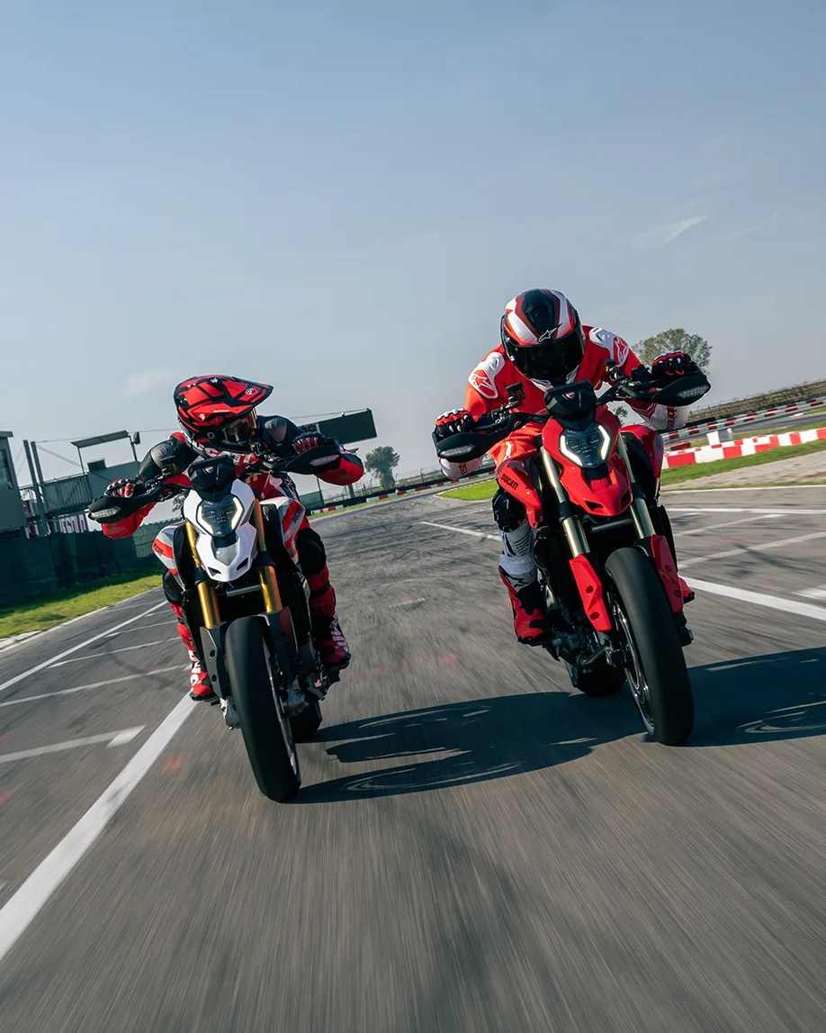Two motorcyclists in red and black gear riding sport motorcycles on a racetrack under clear sky.