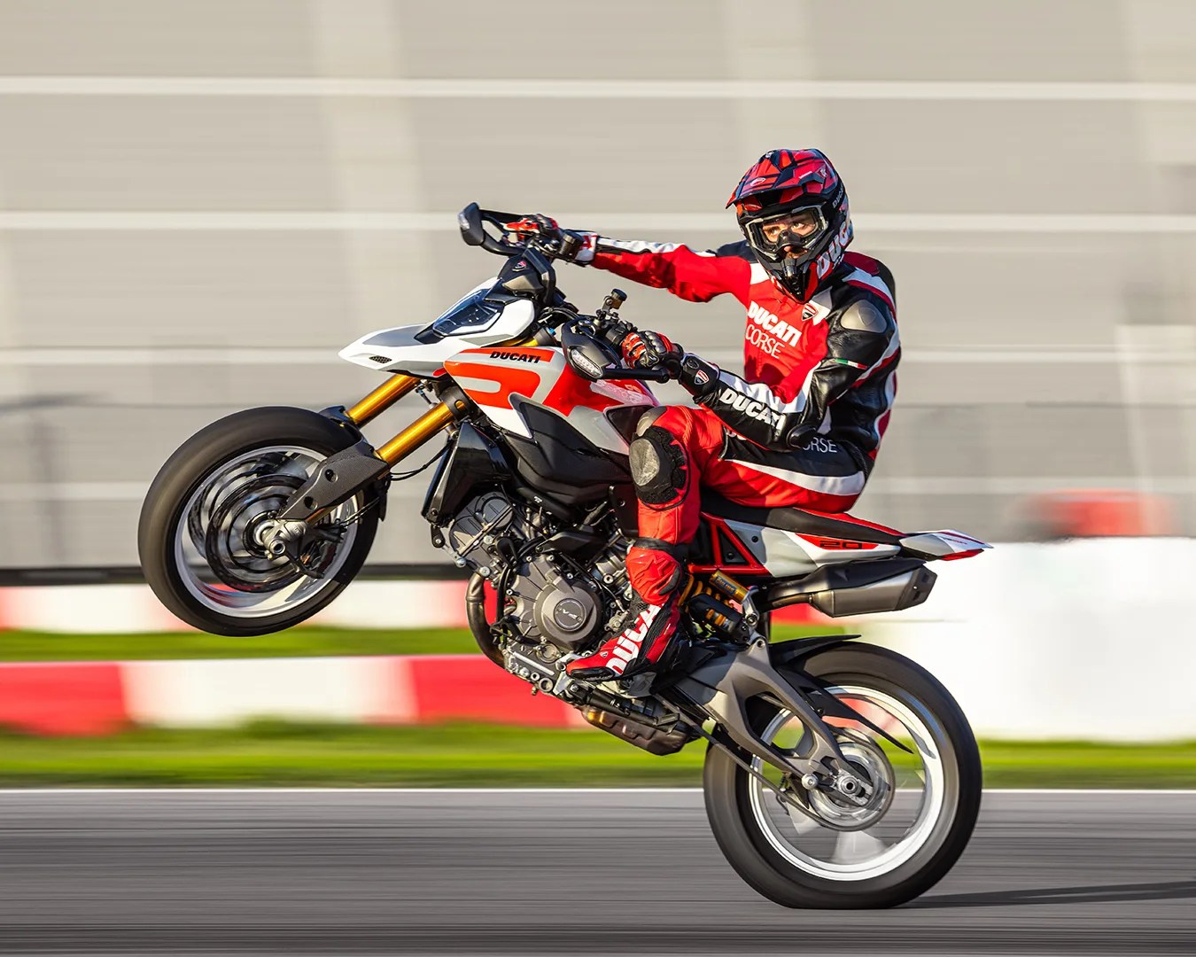 Motorcyclist in red and black Ducati racing gear performing a wheelie on a white and red Ducati motorcycle on a racetrack.