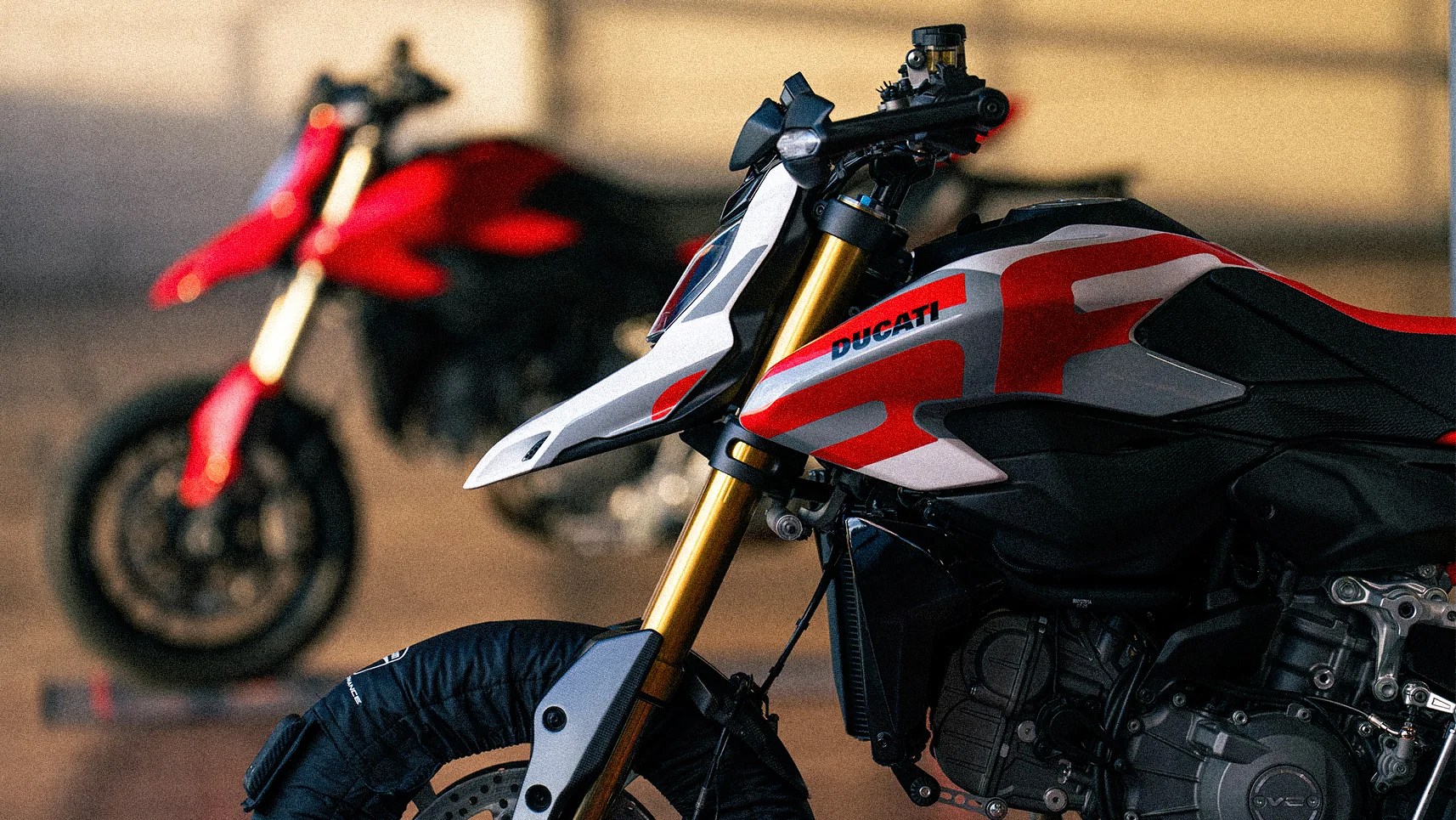 Close-up of a Ducati motorcycle front with gold forks and red, white, and black bodywork, with a blurred red motorcycle in the background.