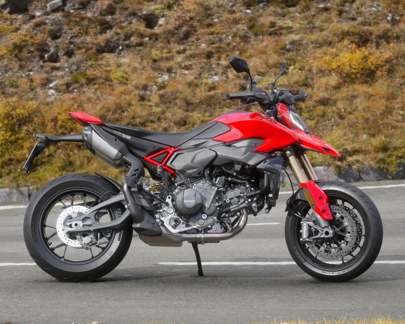 Red and black Ducati motorcycle parked on a road with autumn foliage in the background.