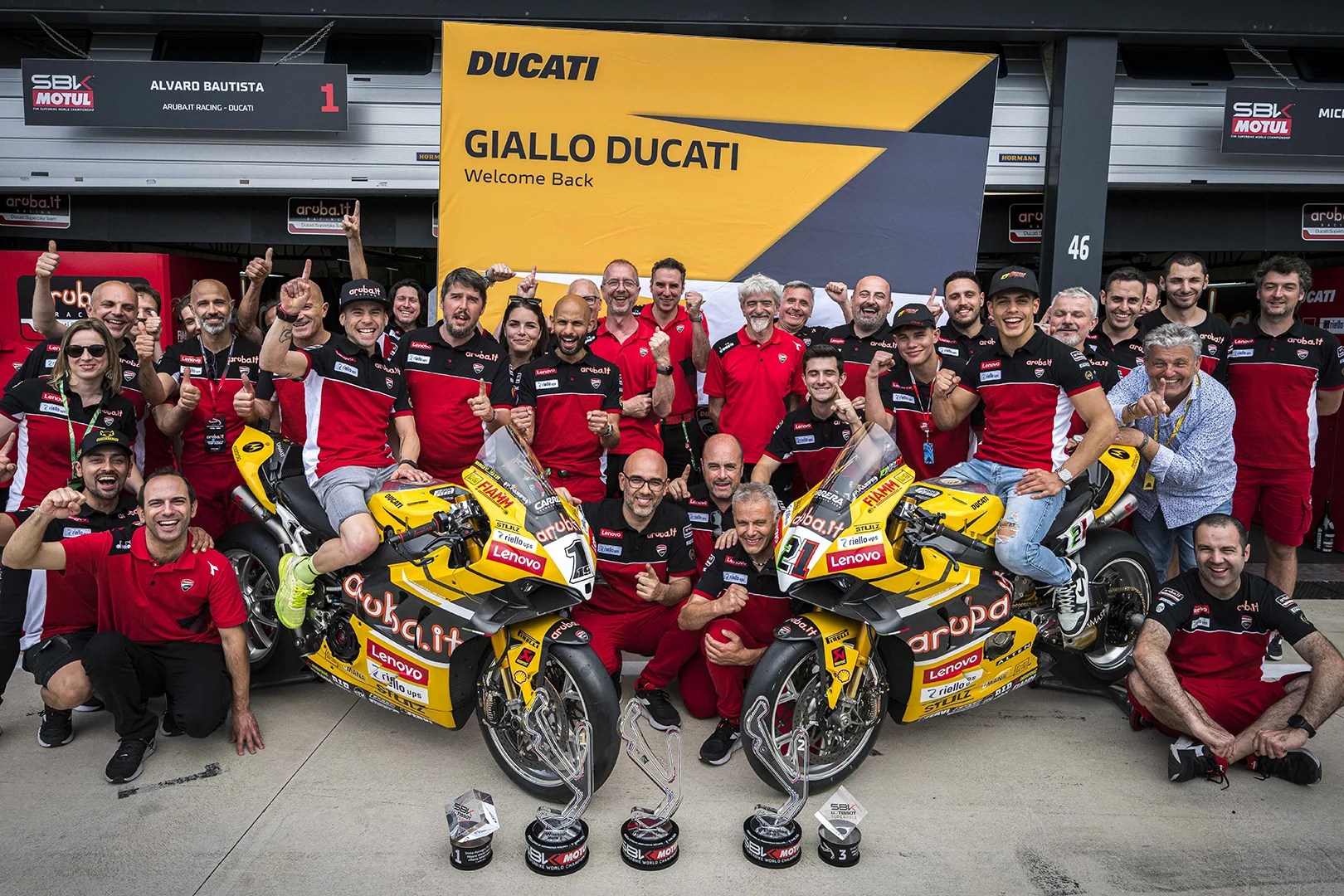 Group of Ducati racing team members posing with two yellow Ducati motorcycles and three trophies in front of a yellow and black "GIALLO DUCATI Welcome Back" banner.