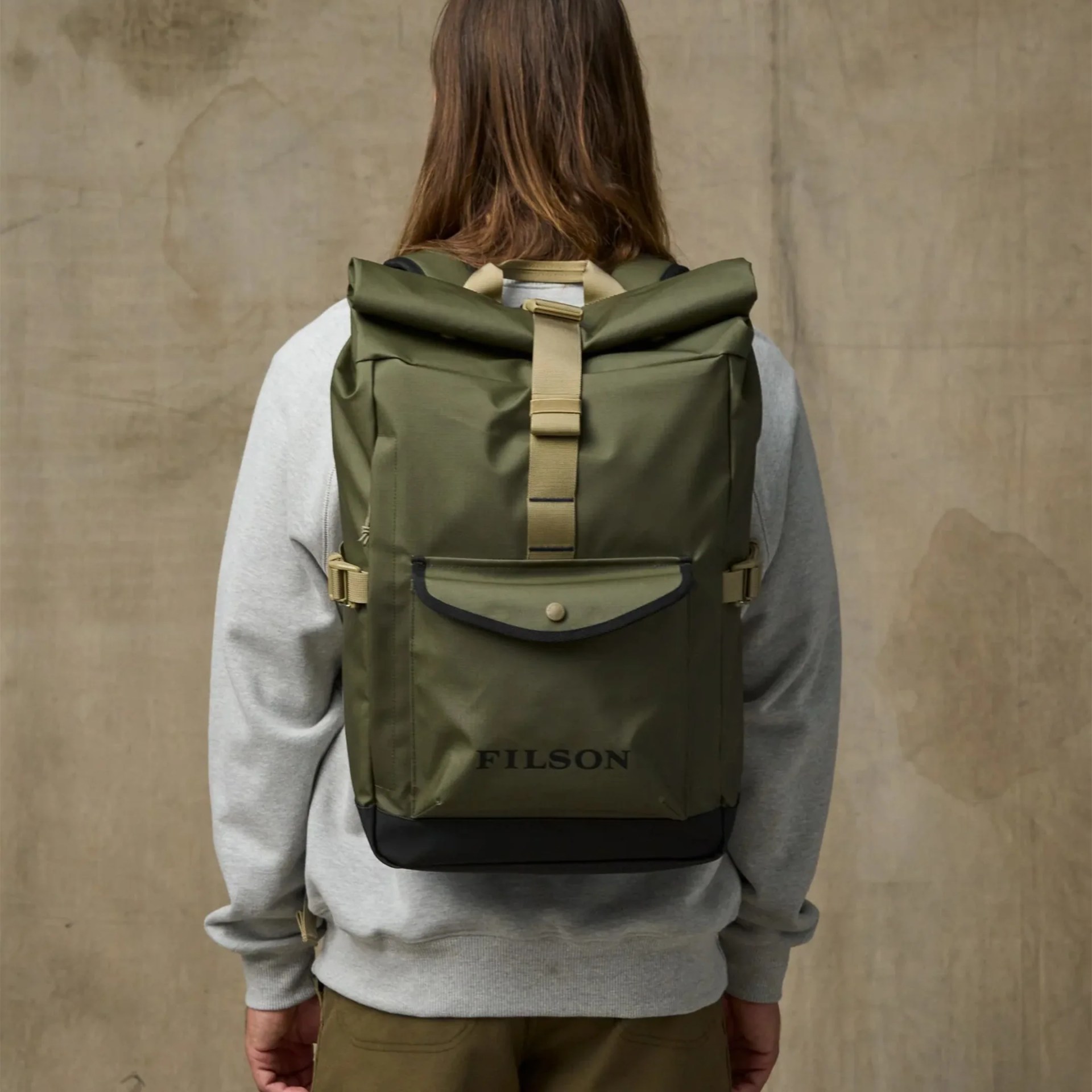 Olive green roll-top backpack with front pocket and beige strap worn by a person in a gray sweatshirt.