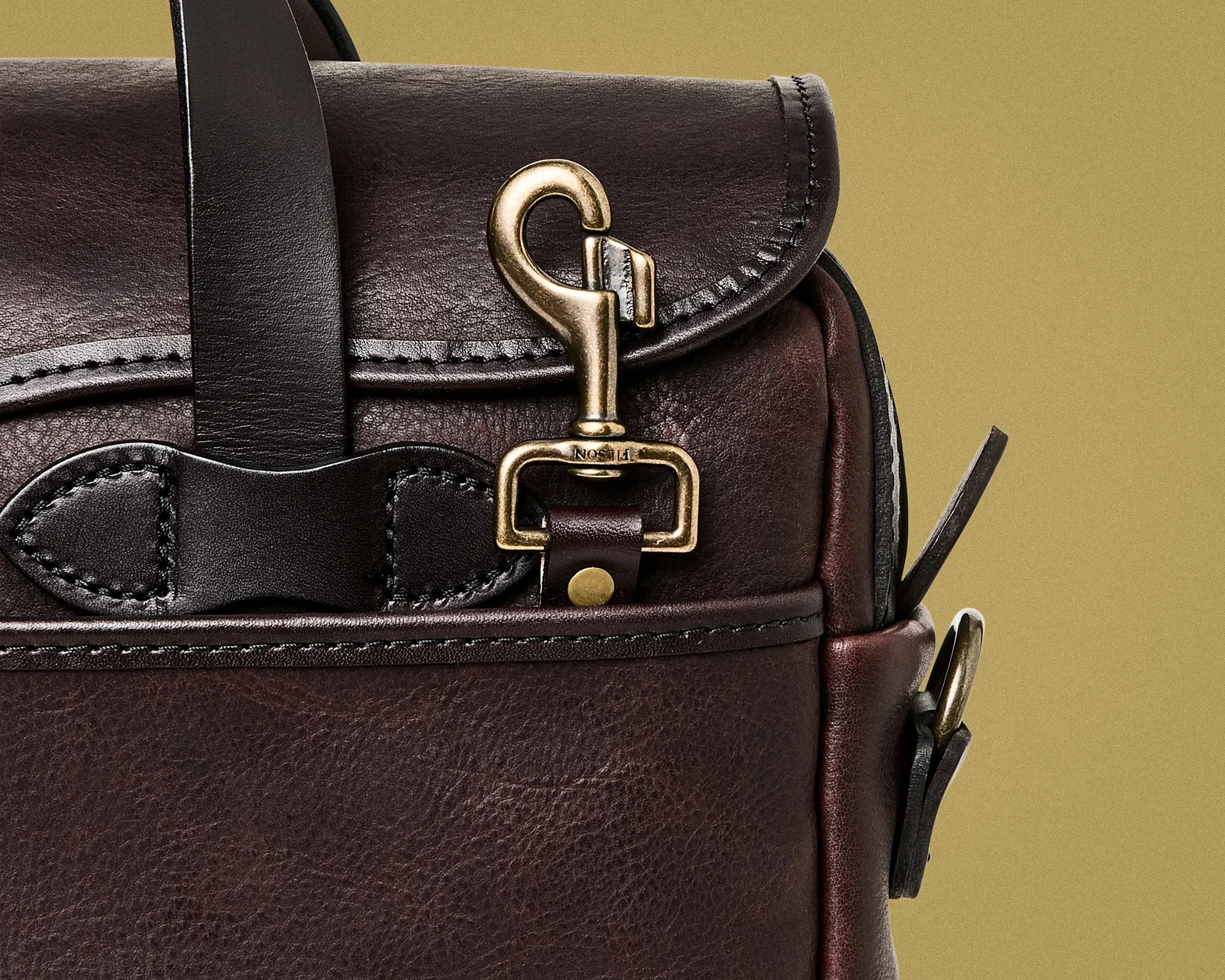Close-up of dark brown leather bag with brass hook clasp and black leather strap detail against a beige background.