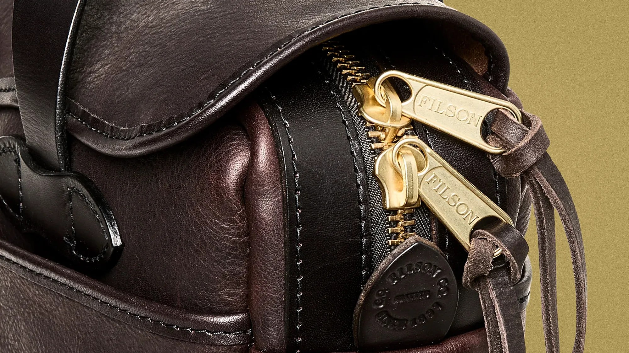 Close-up of dark brown leather bag with two gold zippers engraved "FILSON" and leather zipper pulls.