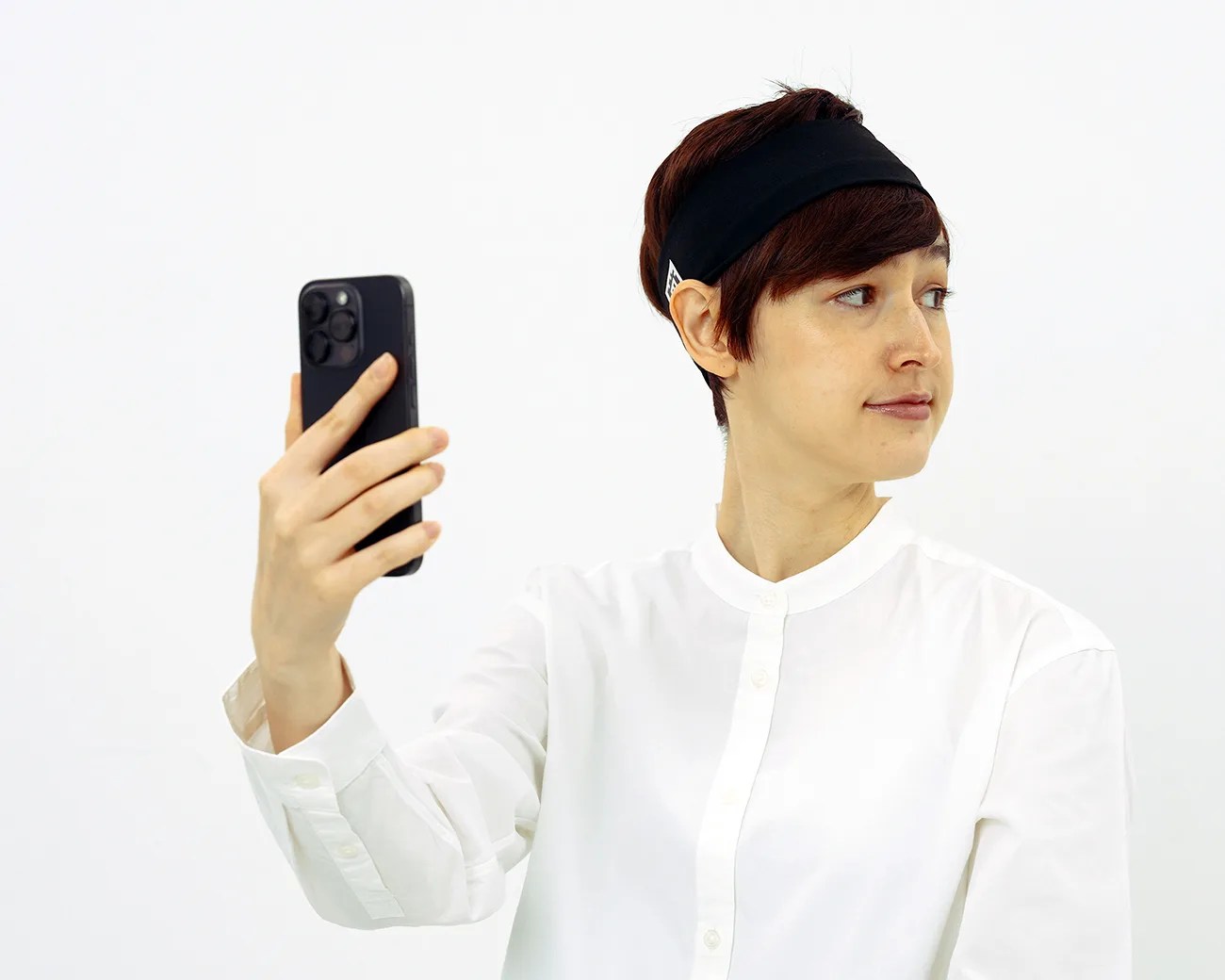 Person in a white button-up shirt and black headband holding a black smartphone in a hand, looking sideways.