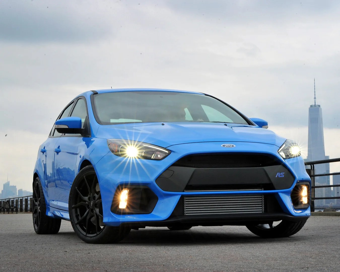 Blue Ford Focus RS hatchback with headlights on parked on a city waterfront with skyscrapers in the background.