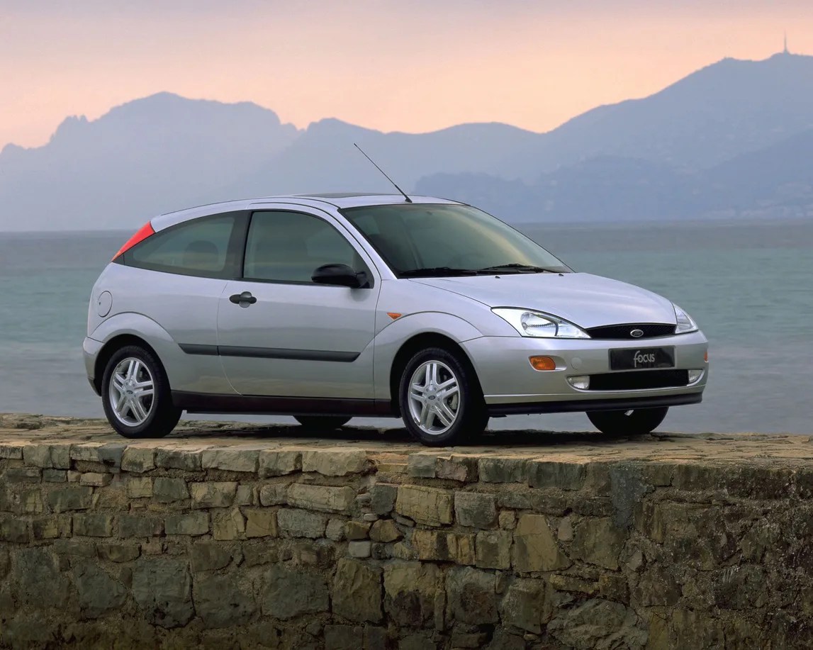 Silver Ford Focus hatchback parked on a stone wall with mountains and sea in the background at sunset.