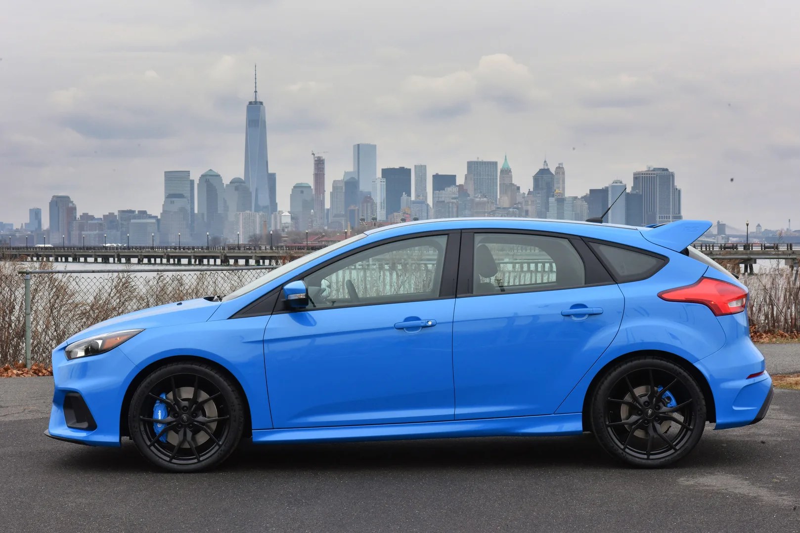Blue sporty hatchback car with black wheels parked on asphalt with a city skyline in the background.